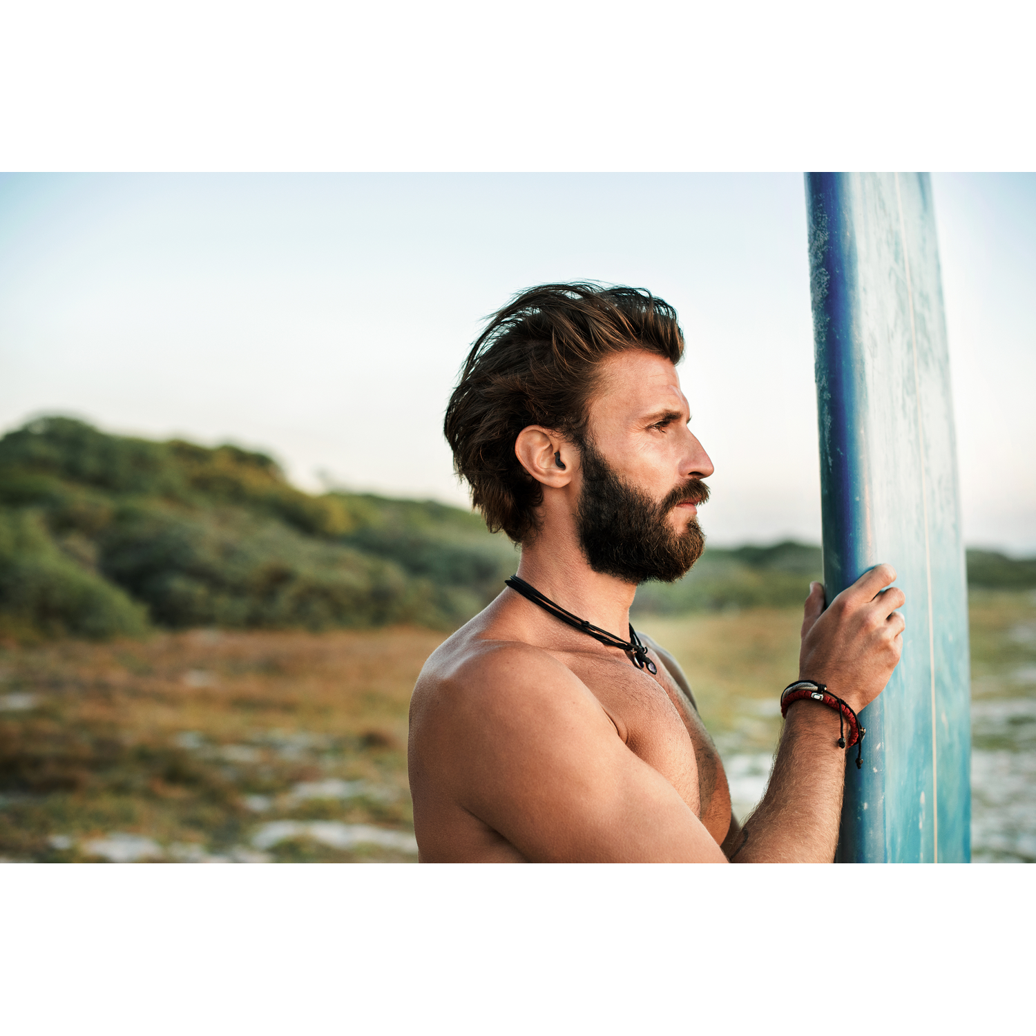 A man with a beard holds a surfboard. Beach and sky in the background.