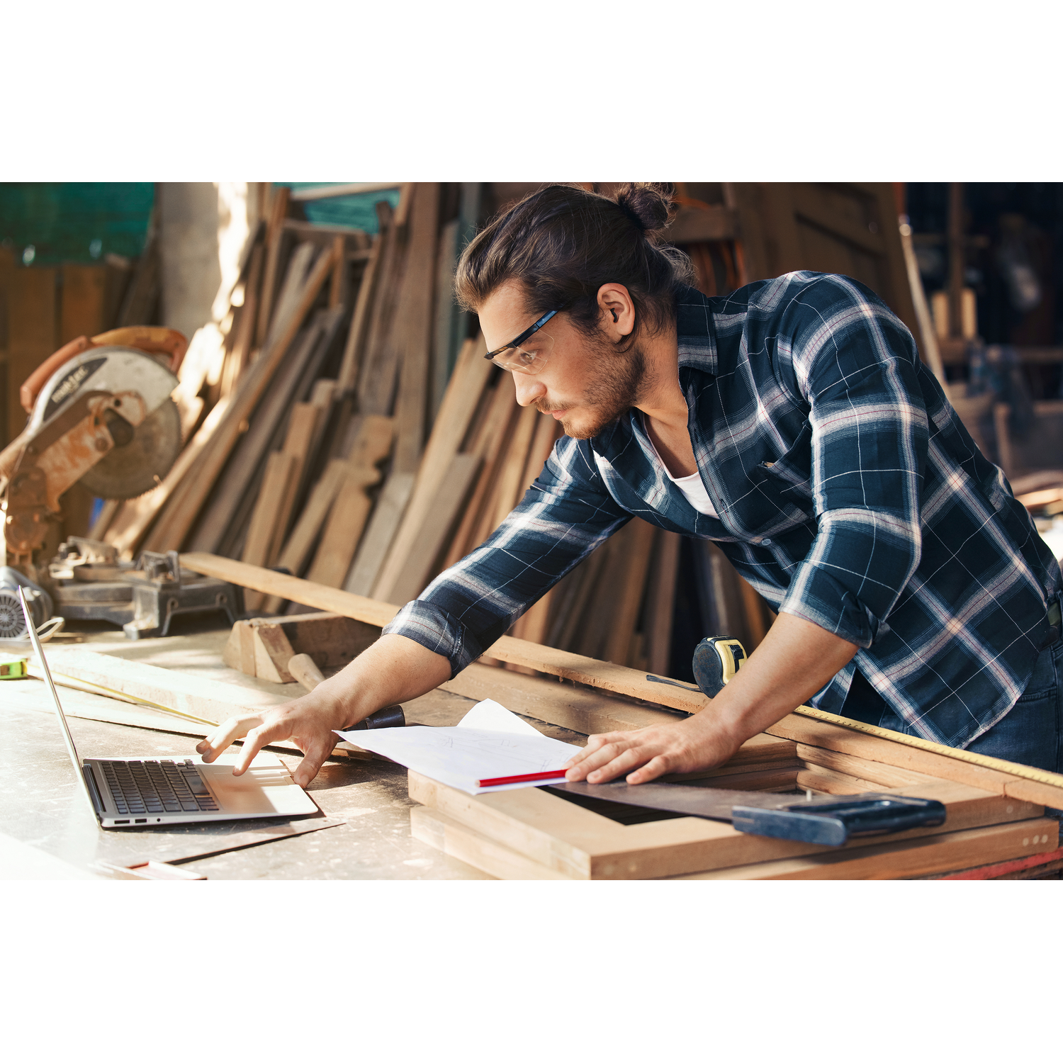 A man working in a workshop, looking at a laptop and plans. He is wearing safety glasses.