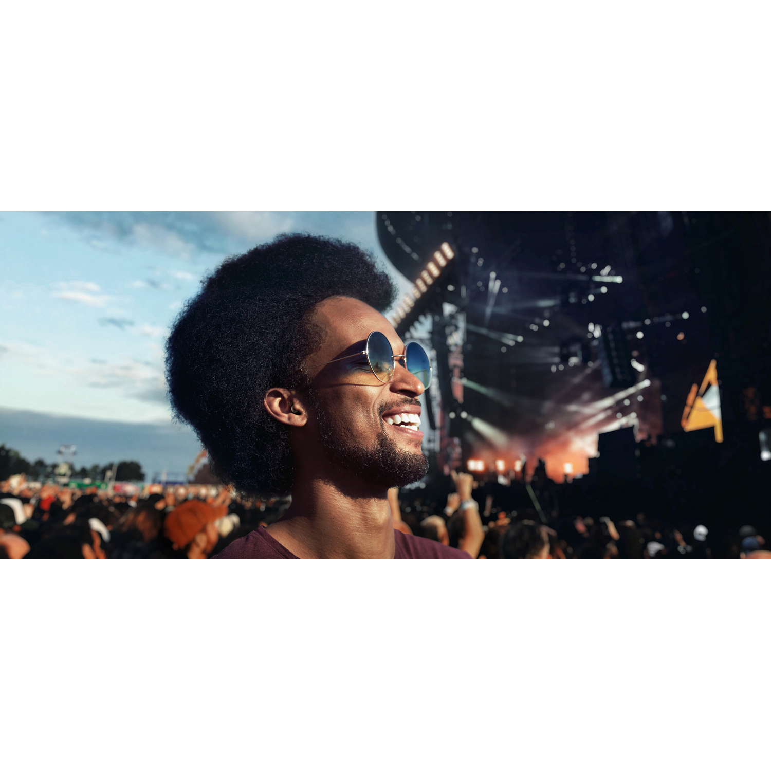 A man with sunglasses smiling at a concert crowd, blue sky in the background.