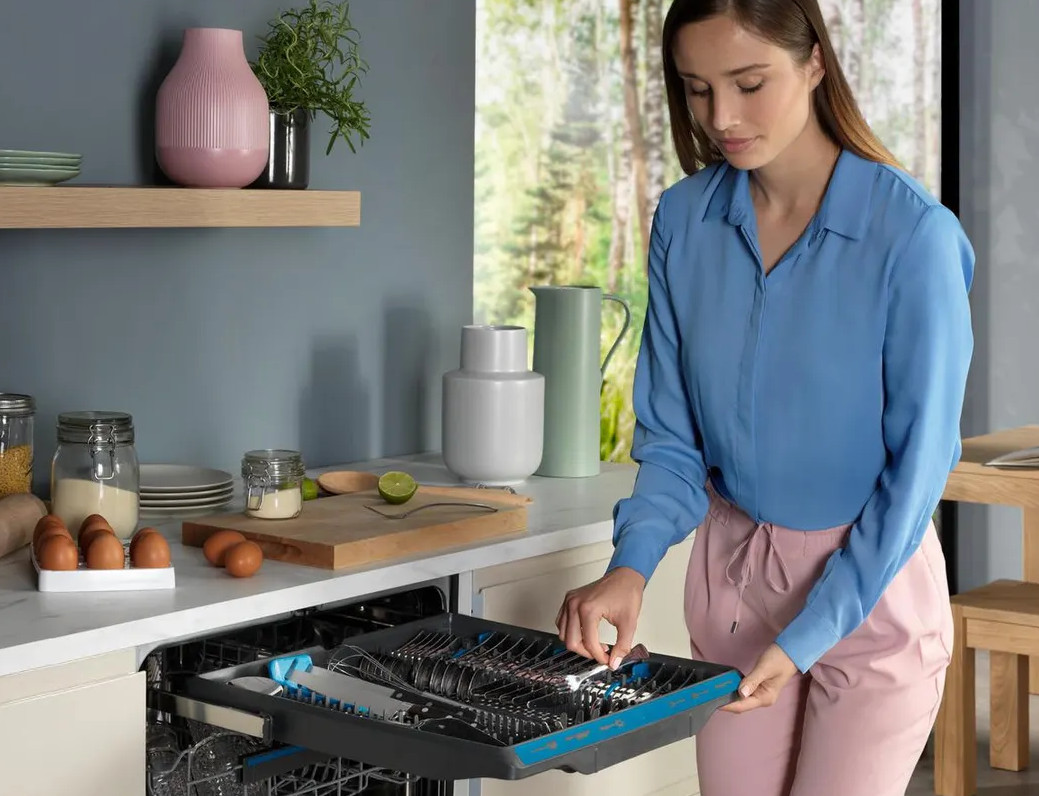 Kobieta załadowuje zmywarkę. Ustawienie kuchenne z naczyniami, przyborami i niebieską zmywarką.