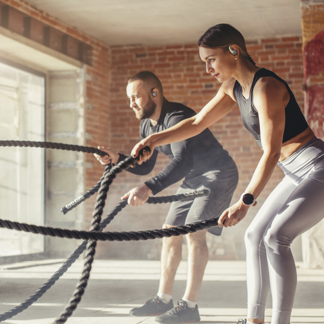 Zwei Personen trainieren mit Seilen in einem Fitnessstudio mit Backsteinmauer.