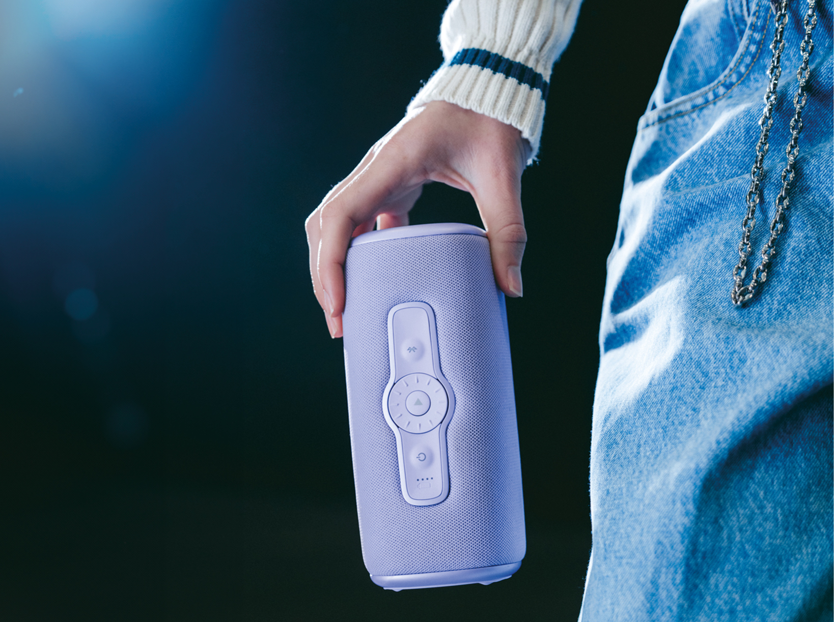 A person holds a lavender purple speaker with buttons. Black background with a blue light.