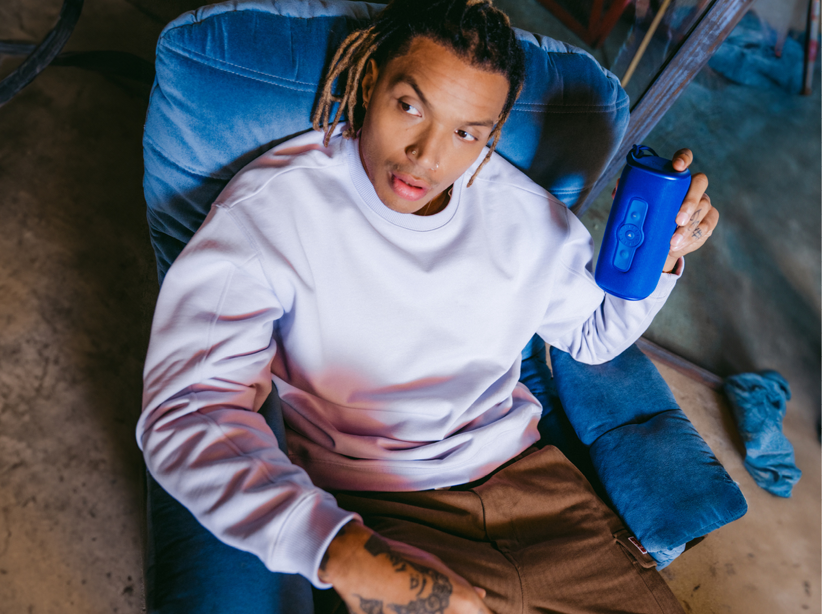 A man sits in a blue velvet chair holding a blue speaker.