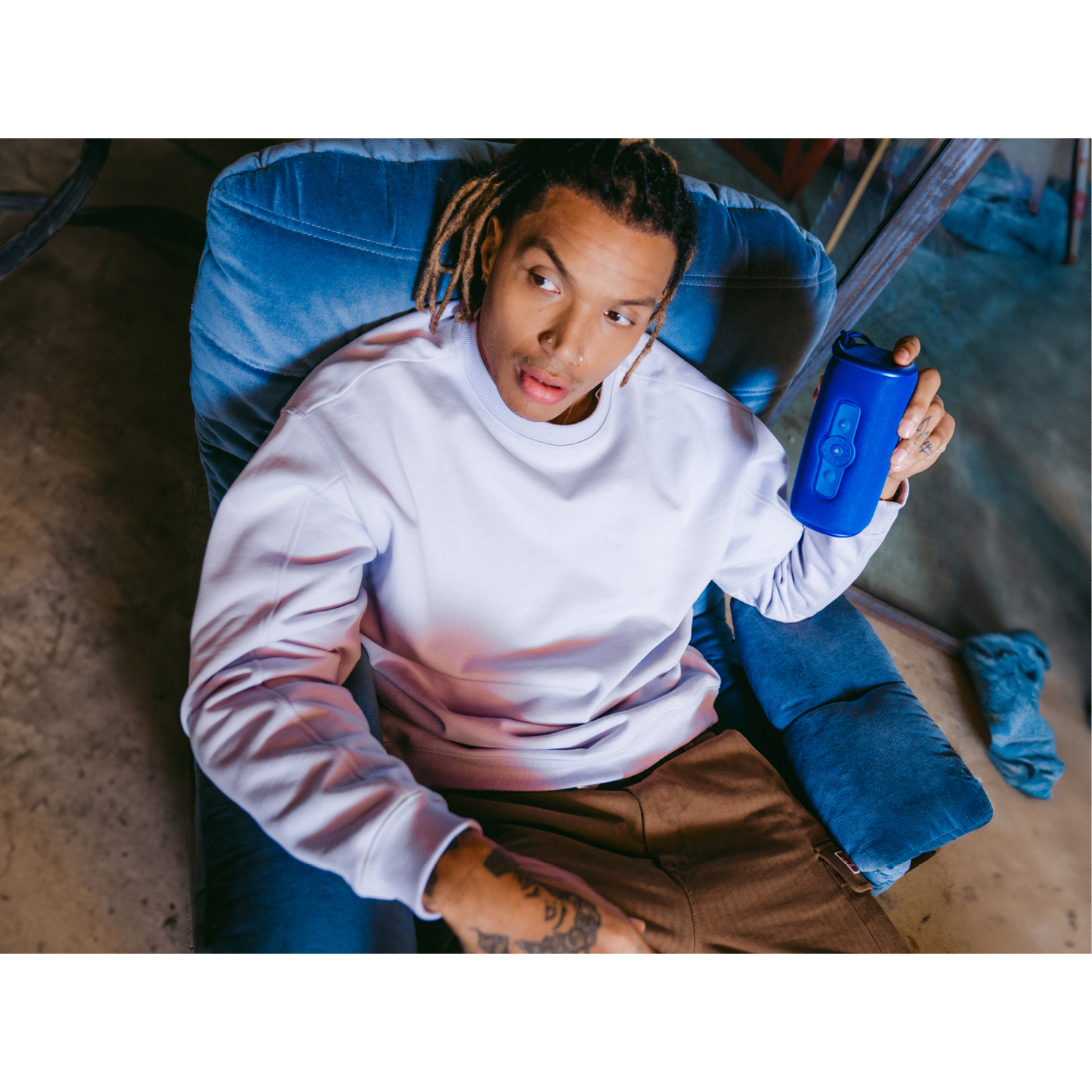 A man with dreadlocks sits in a blue chair, holding a blue bottle. He looks upwards.