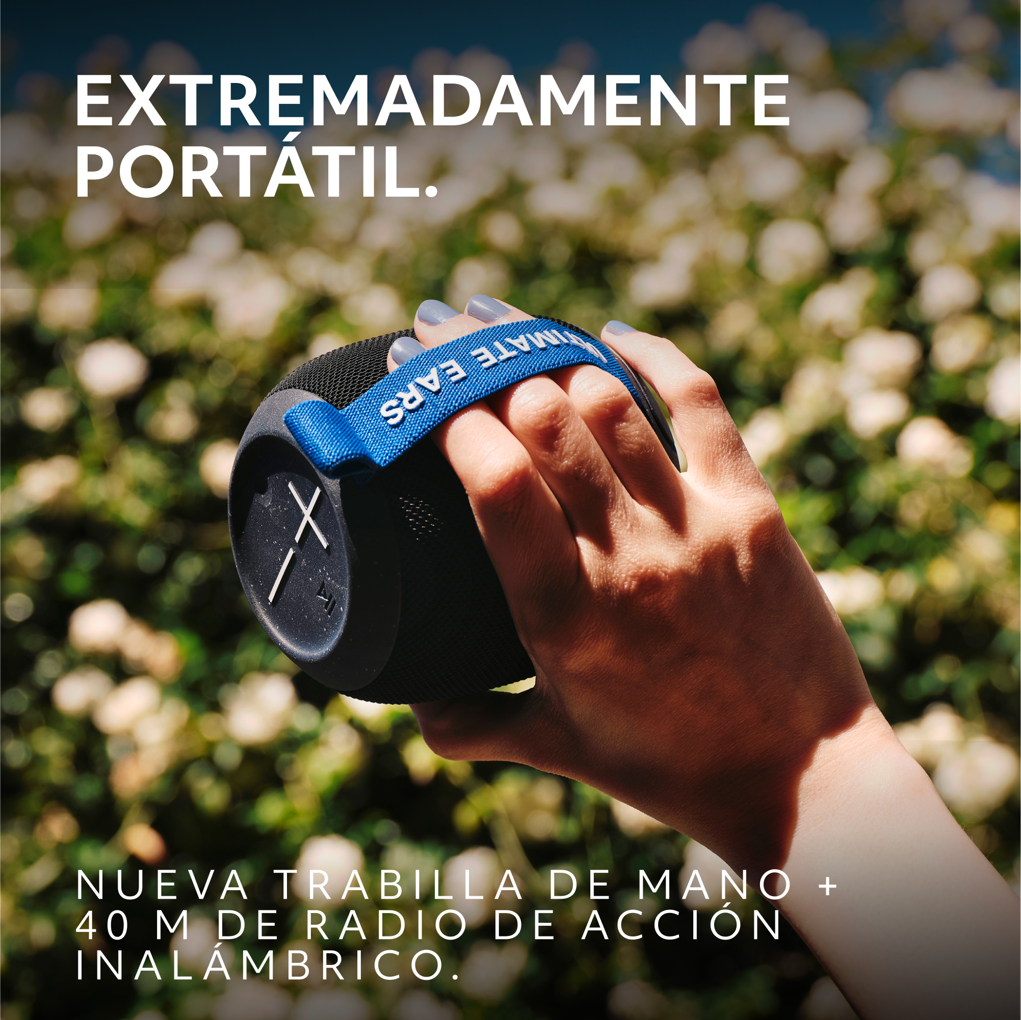 Una mano sostiene un altavoz portátil negro y azul frente a un fondo floral borroso verde y blanco.