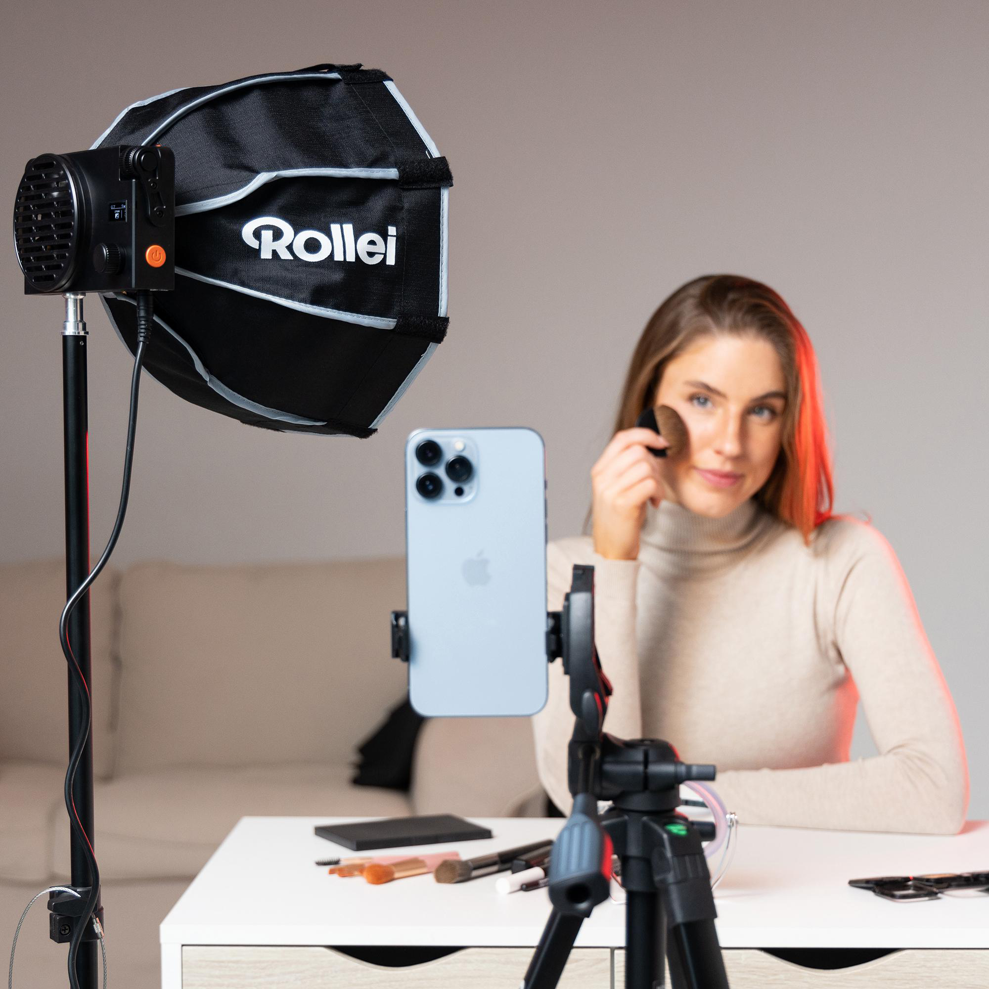 Frau schminkt sich mit Telefon auf Stativ. Ringlicht und Make-up-Produkte auf dem Tisch.