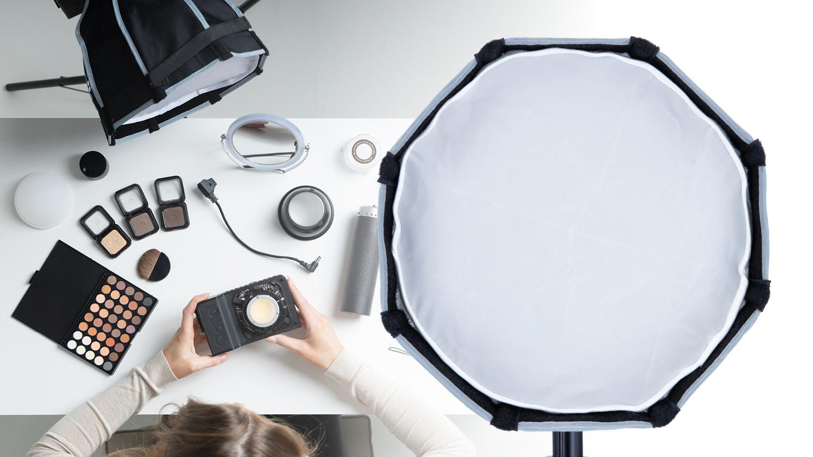 Draufsicht auf Make-up und Beleuchtungsausrüstung auf einem weißen Tisch, Hände halten ein Gerät.