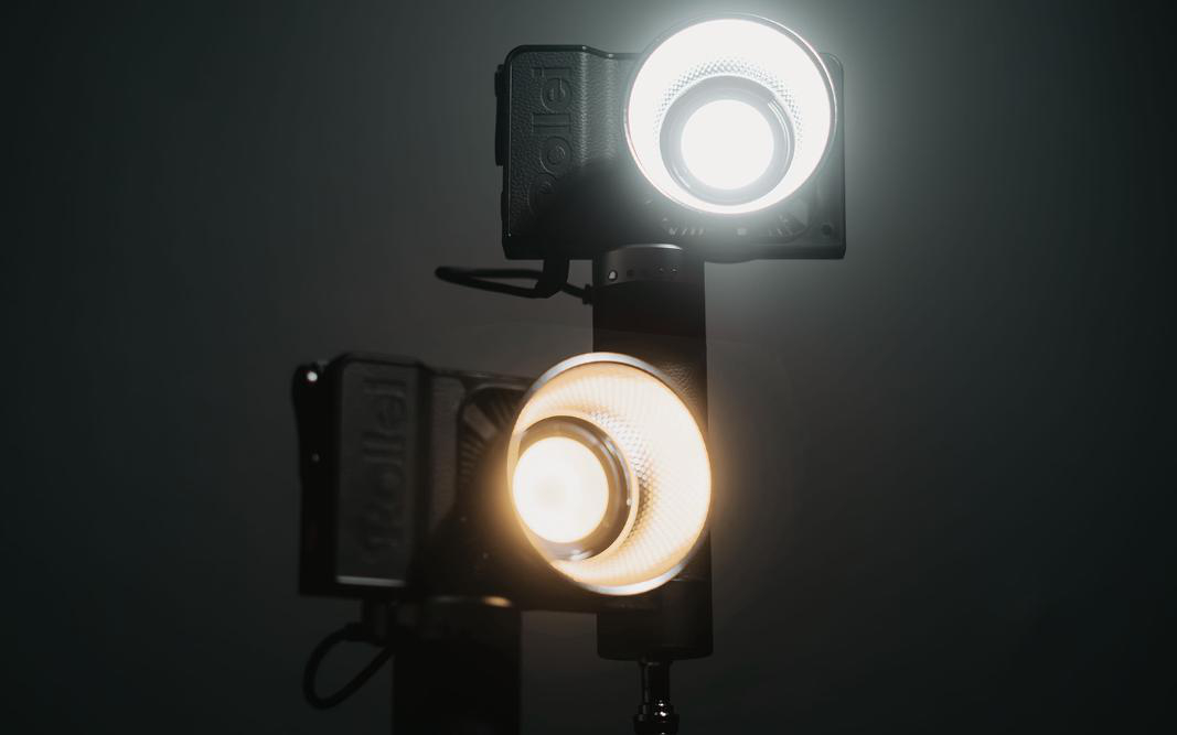 Zwei schwarze Rollei-Leuchten auf einem Ständer, beleuchtet. Dunkler Hintergrund.