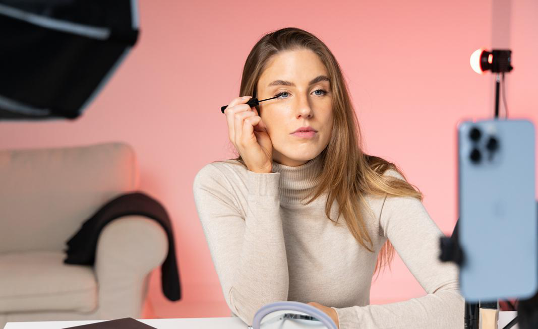 Frau trägt Mascara auf, während sie mit dem Telefon filmt. Rosa Hintergrund.