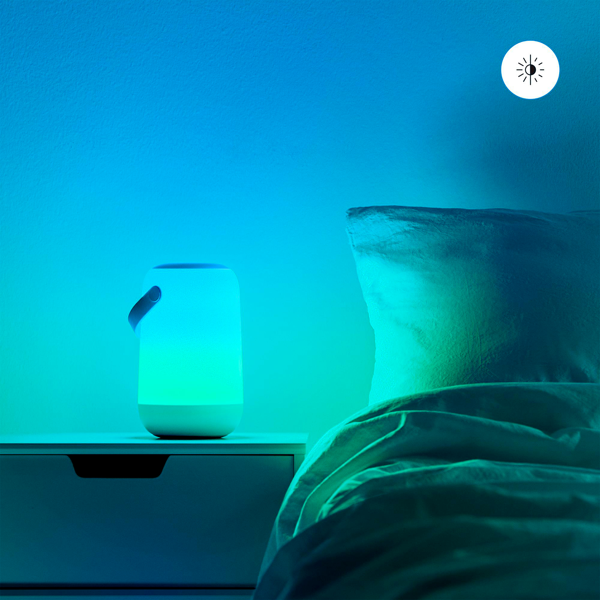 Eine leuchtende Lampe auf einem Nachttisch neben einem Bett. Das Licht spiegelt sich an Wand und Bettwäsche.