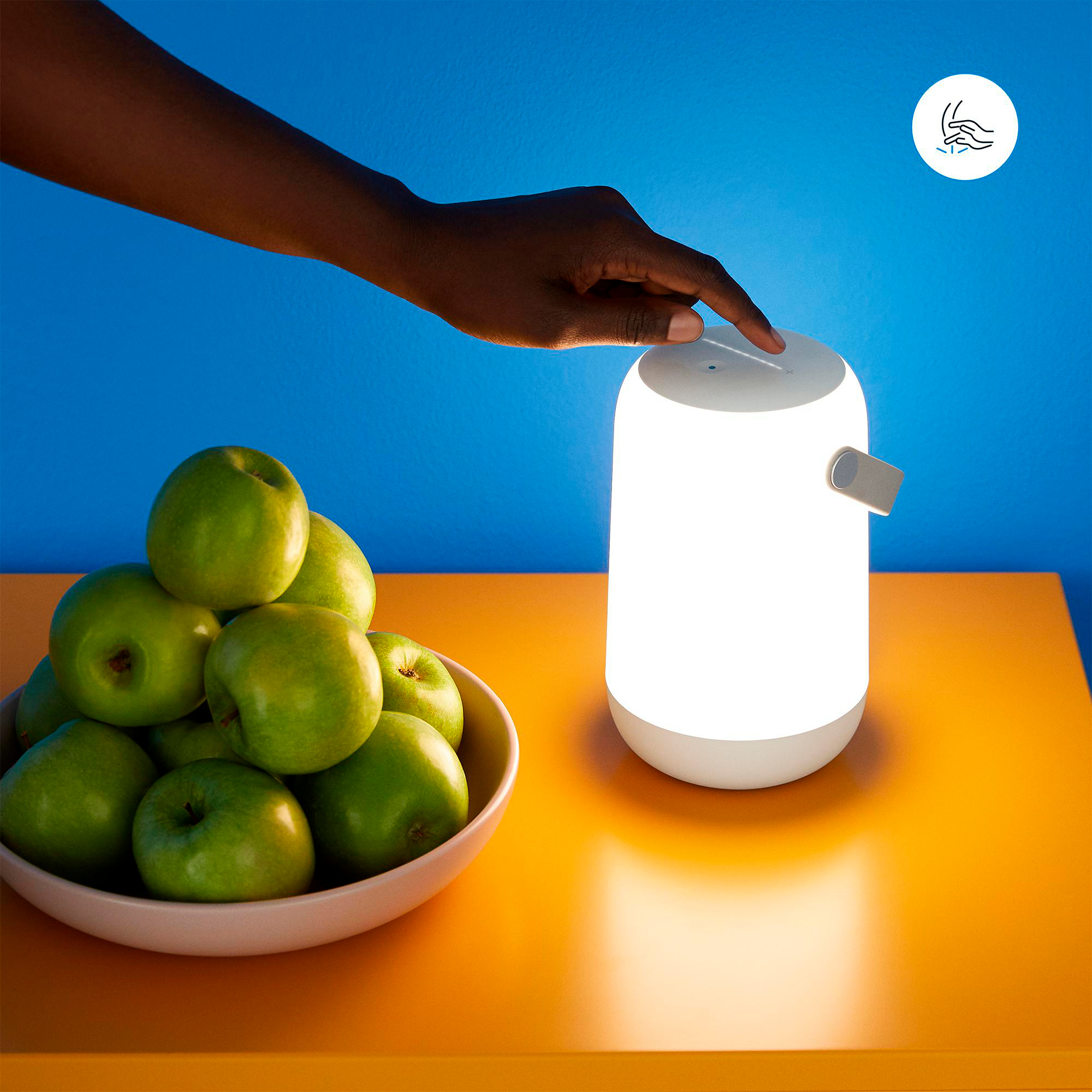 Eine Hand berührt eine weiße Lampe auf einem orangefarbenen Tisch. Grüne Äpfel sind in einer Schüssel. Blauer Hintergrund.