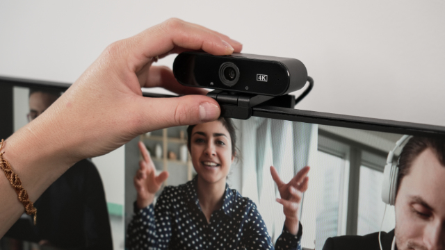 Egy kéz állít be egy fekete webkamerát, ami egy videohívást megjelenítő monitorra van rögzítve.