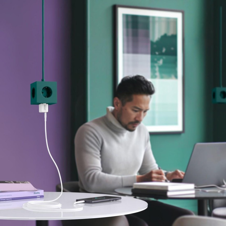 Mężczyzna pracuje na laptopie, listwa zasilająca, biały kabel, fioletowa ściana, zielona ściana, stół.