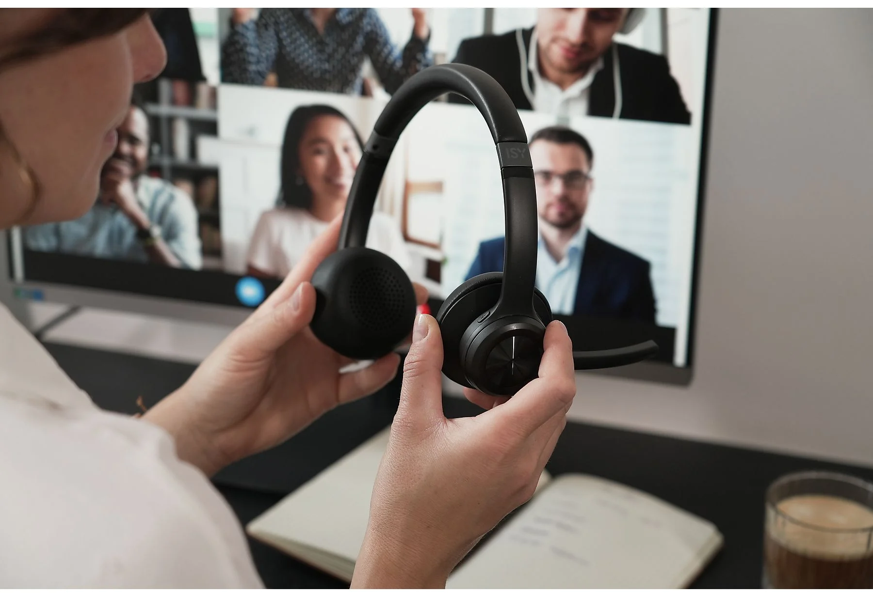 Egy nő fekete fejhallgatót tart egy monitoron lévő videokonferencia előtt.