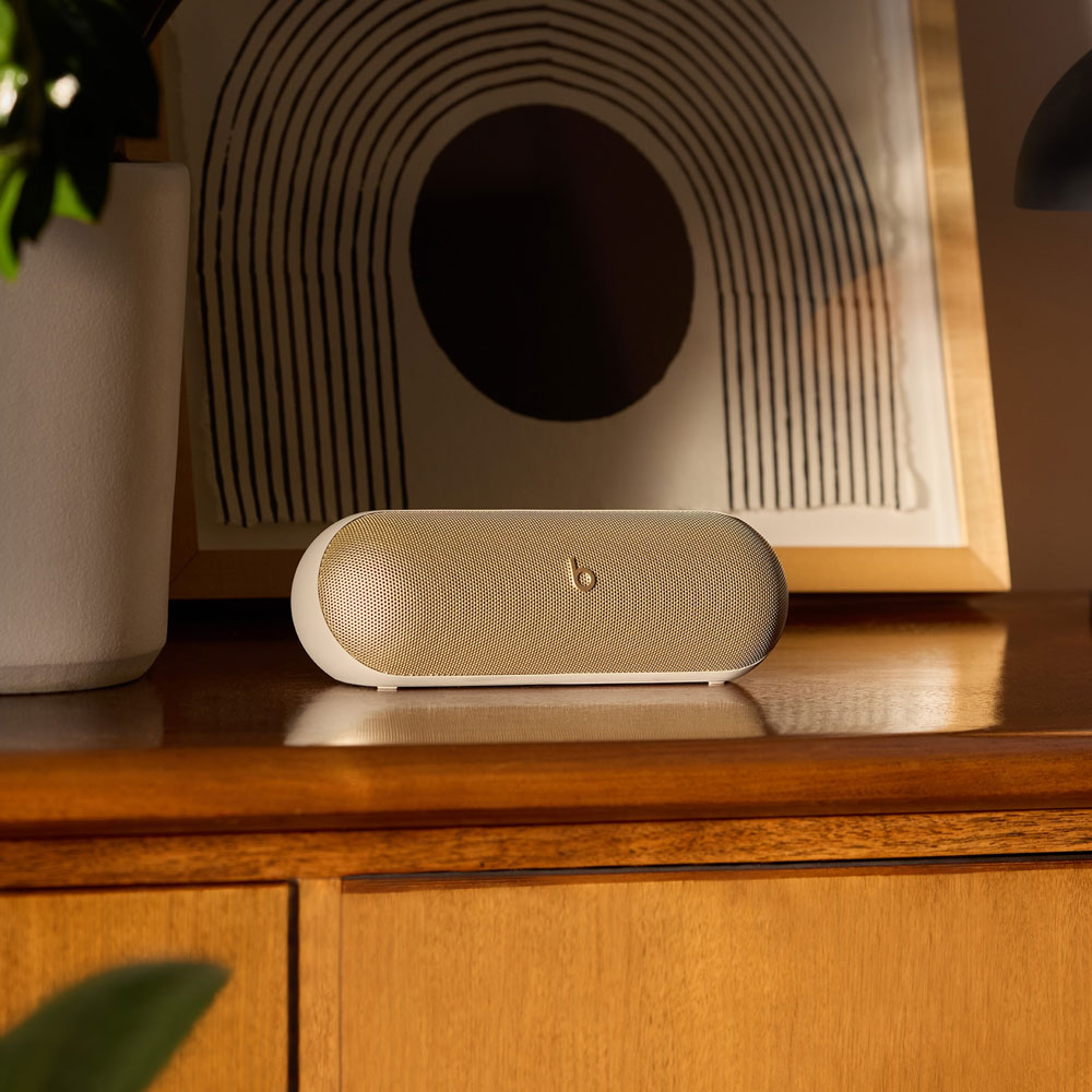 Haut-parleur Beats blanc et doré sur une surface en bois, contre une œuvre encadrée.