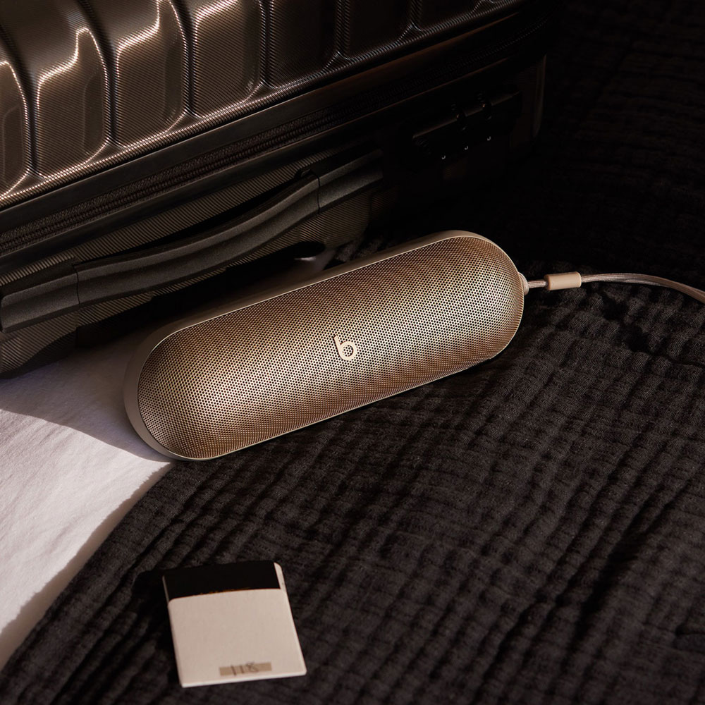 Un haut-parleur Beats doré est connecté à une valise avec un câble. Une petite carte blanche est sur le lit.