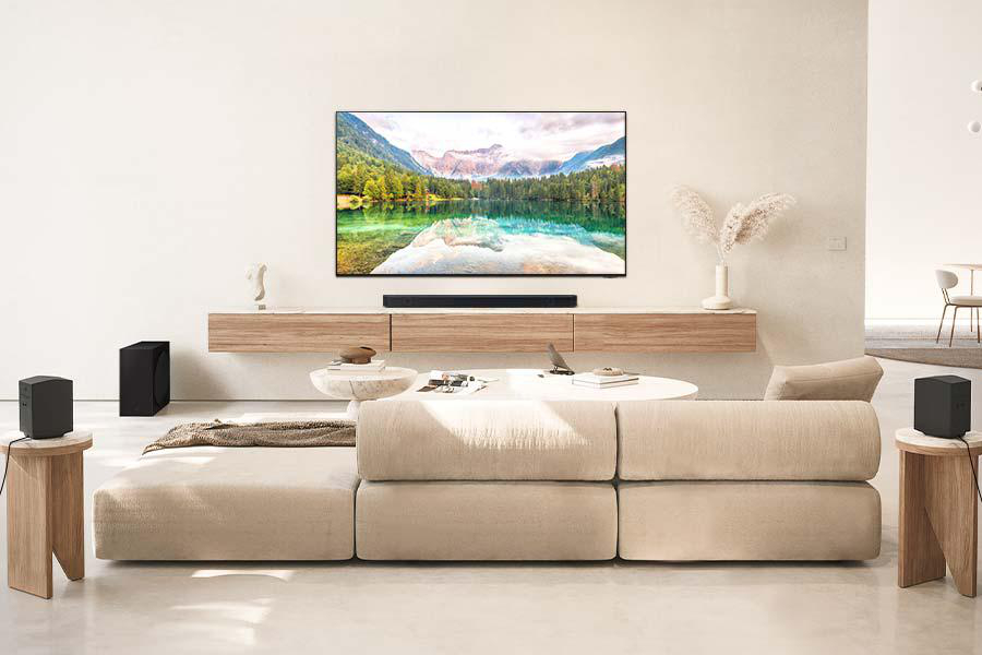 Wohnzimmer-Setup mit großem Fernseher, Soundbar und Lautsprechern auf beigem Sofa.