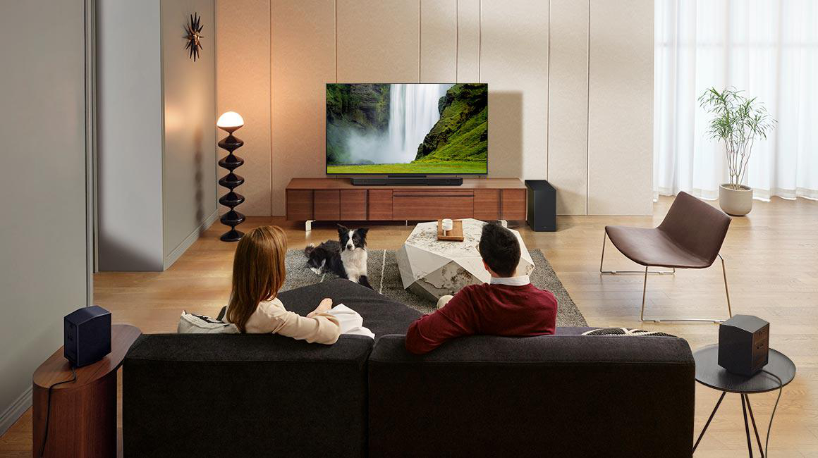 Ein Wohnzimmer mit Fernseher, einem Paar auf dem Sofa und einem Hund. Lautsprecher sind sichtbar.