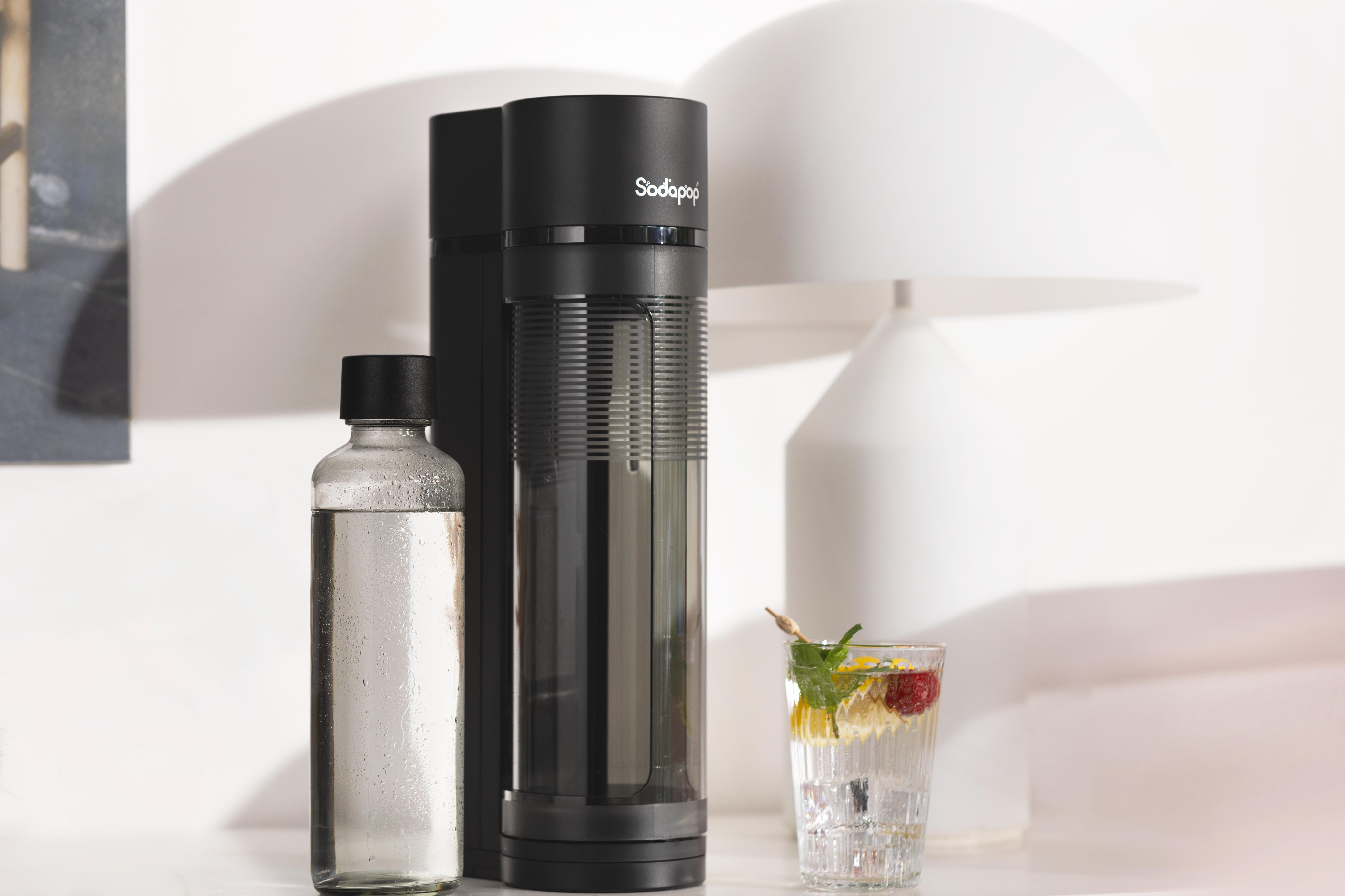 Schwarze SodaPop-Maschine und -Flasche. Ein Glas mit Eis, Zitrone und einer roten Frucht. Weißer Hintergrund.