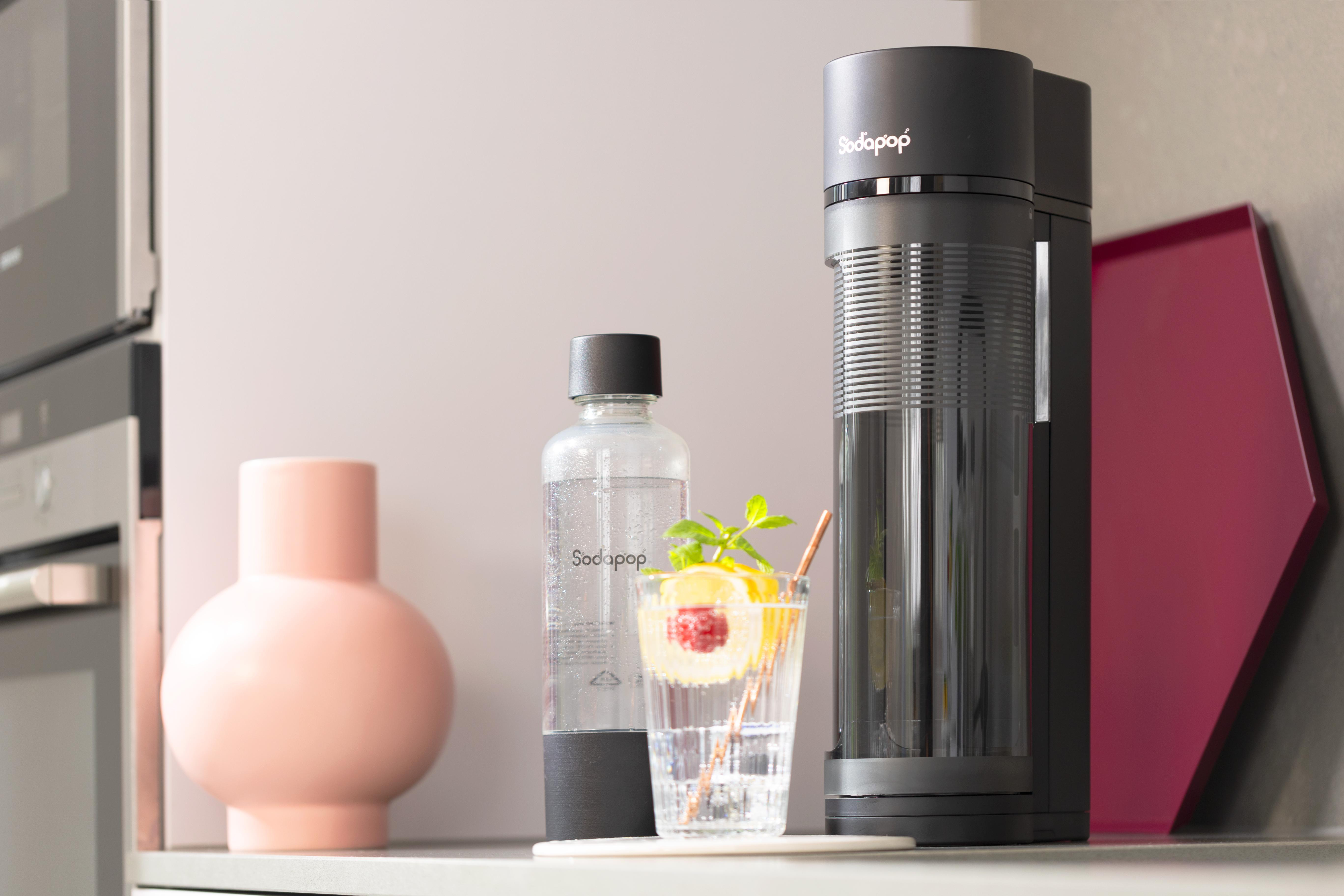 Ein Sodabereiter, eine Flasche und ein Glas mit Strohhalm und Zitronenscheibe stehen auf einer Theke.