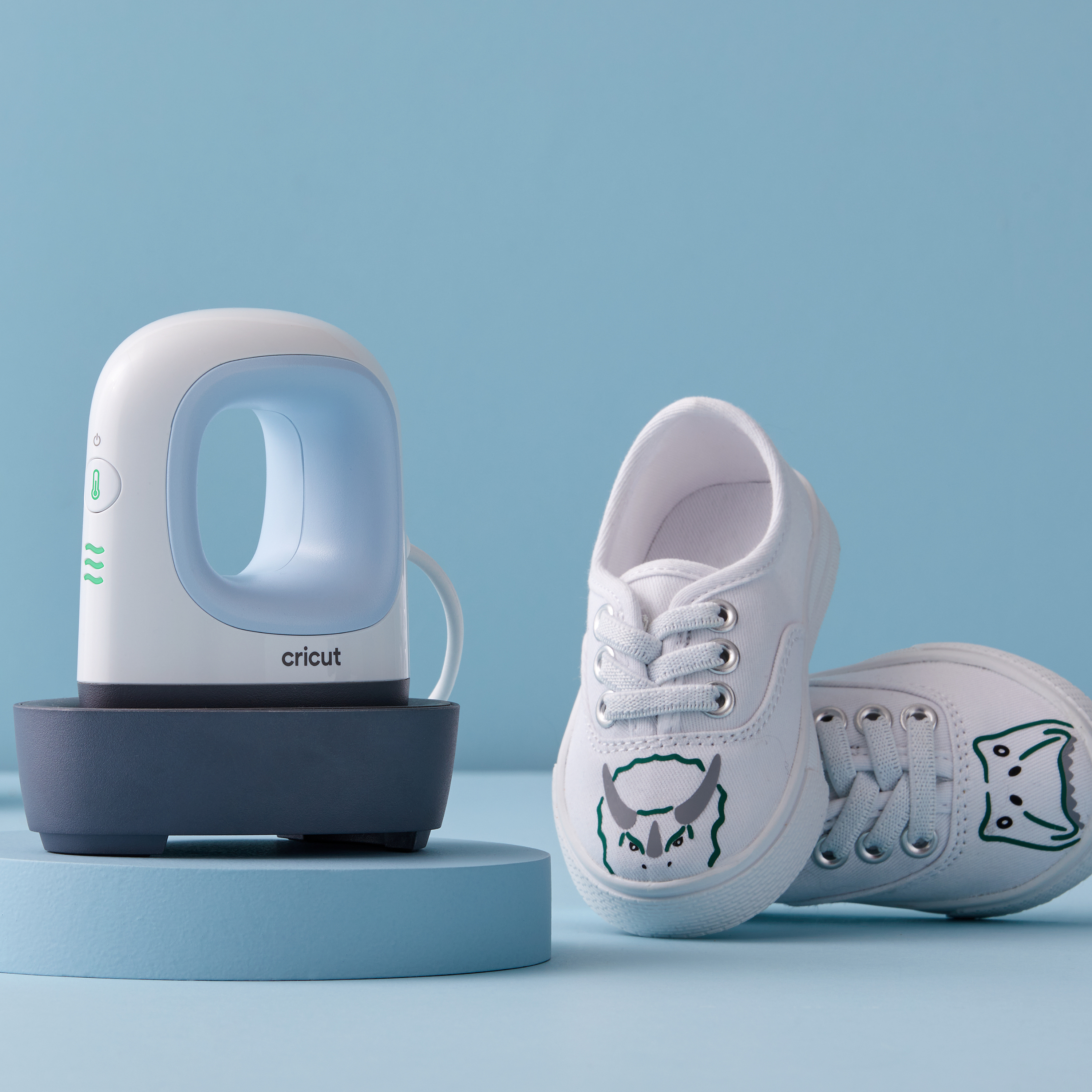 Cricut-Hitzepresse und zwei weiße Schuhe mit Dinosaurier-Design auf blauem Ständer.