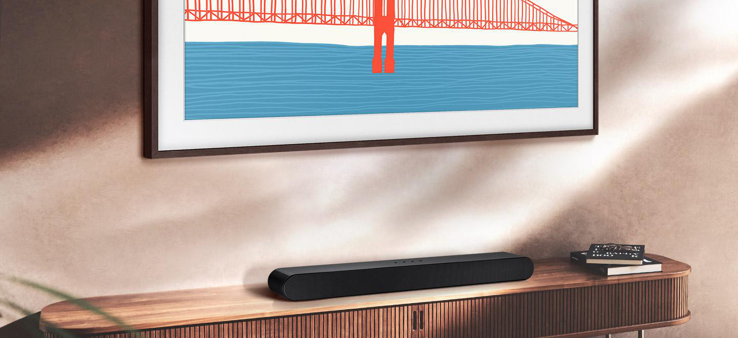 Schwarze Soundbar auf einer Holzkonsol. Im Hintergrund ein Fernseher mit einem Golden-Gate-Bridge-Gemälde.