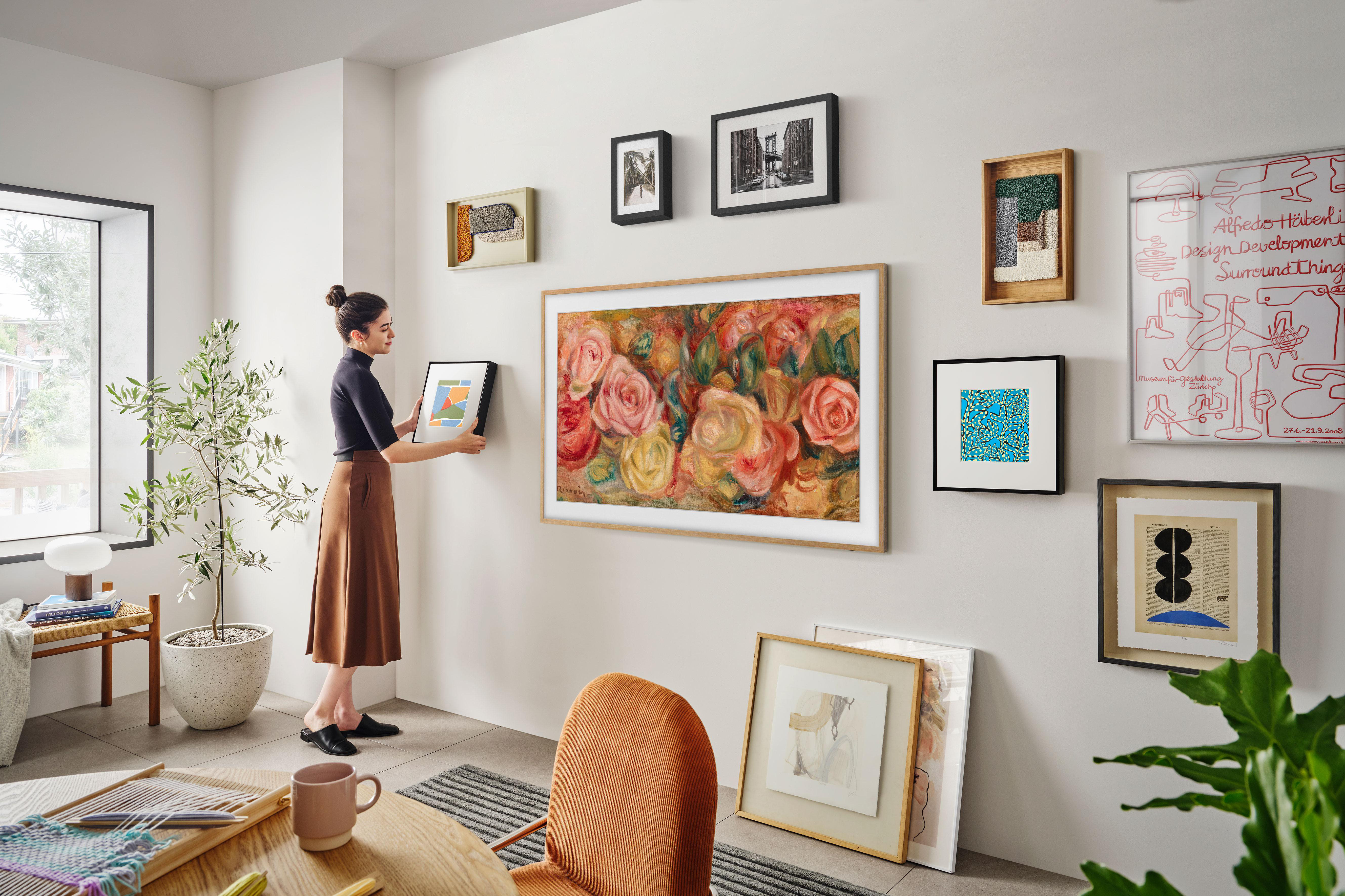 Frau hängt ein gerahmtes Gemälde an einer Wand mit anderen gerahmten Kunstwerken und einem großen Blumengemälde.