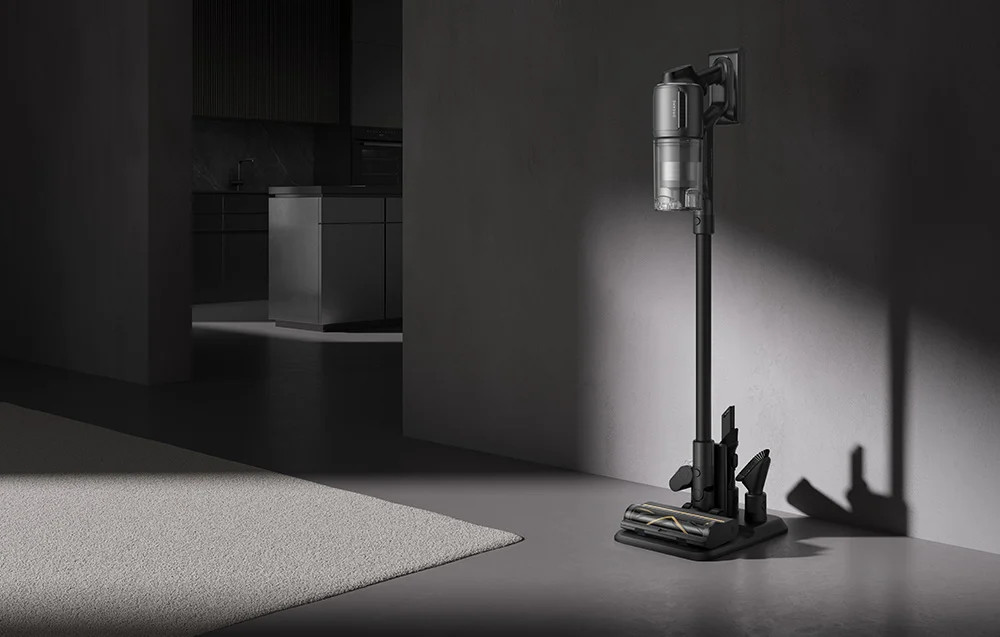 Czarny odkurzacz ze stojakiem w pokoju. Szare ściany, biały dywan. Kuchnia widoczna w tle.