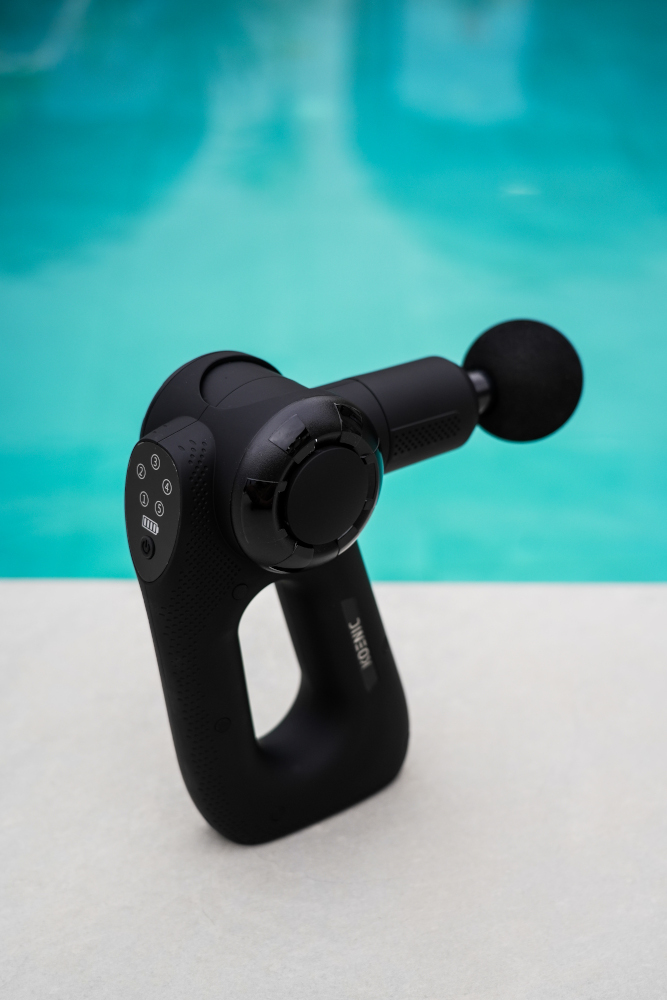 Black massage gun with a round head, on a white surface, pool in the background.