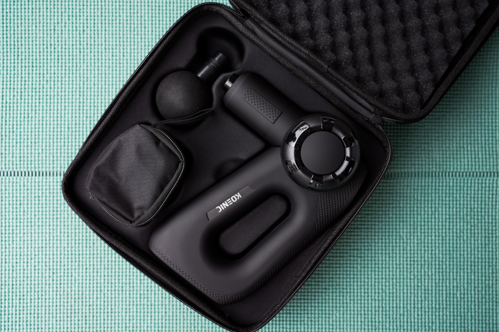 Black massage gun in a case, with accessories. On a green textured surface.