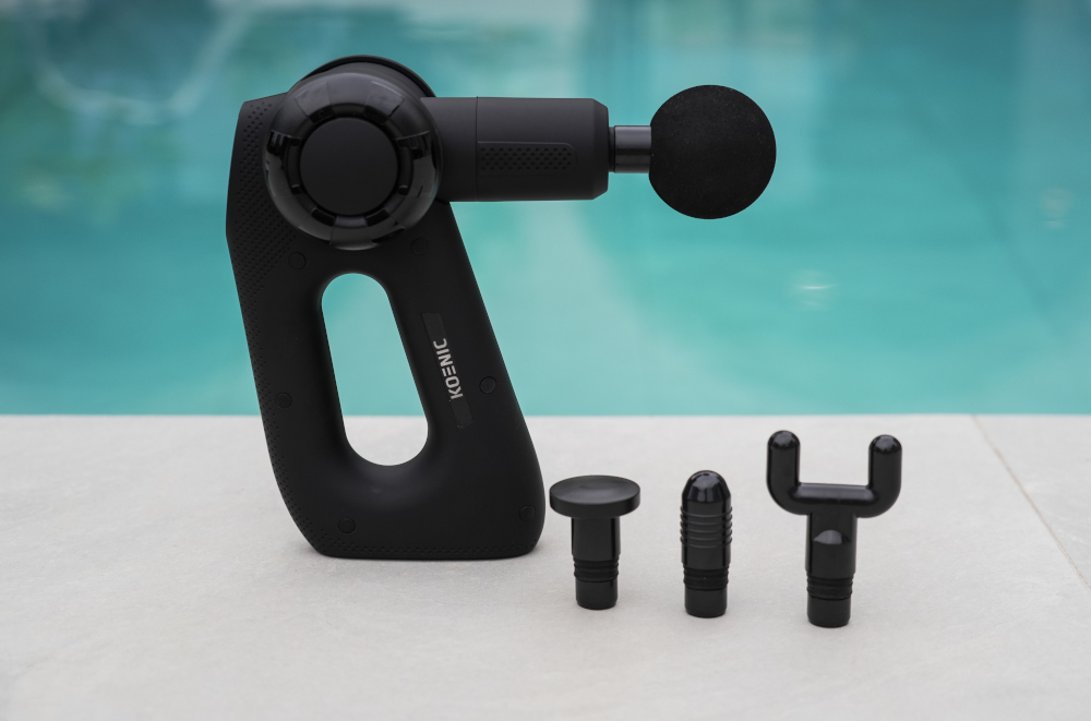 Black massage gun with multiple attachments on a white surface, water background.