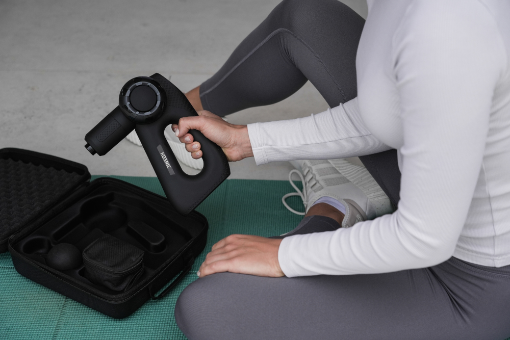 A person is holding a black massage gun, with the case open and accessories visible.