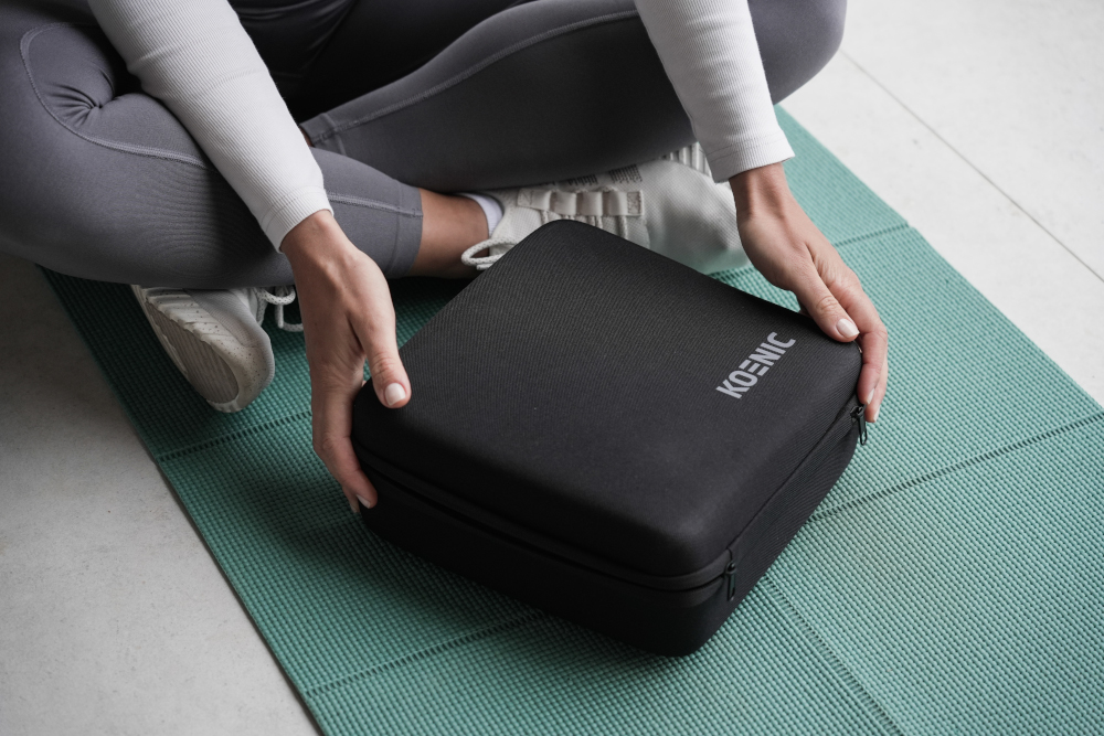 A person sitting on a yoga mat, holding a black square case with the word 'KOENIC' on it.