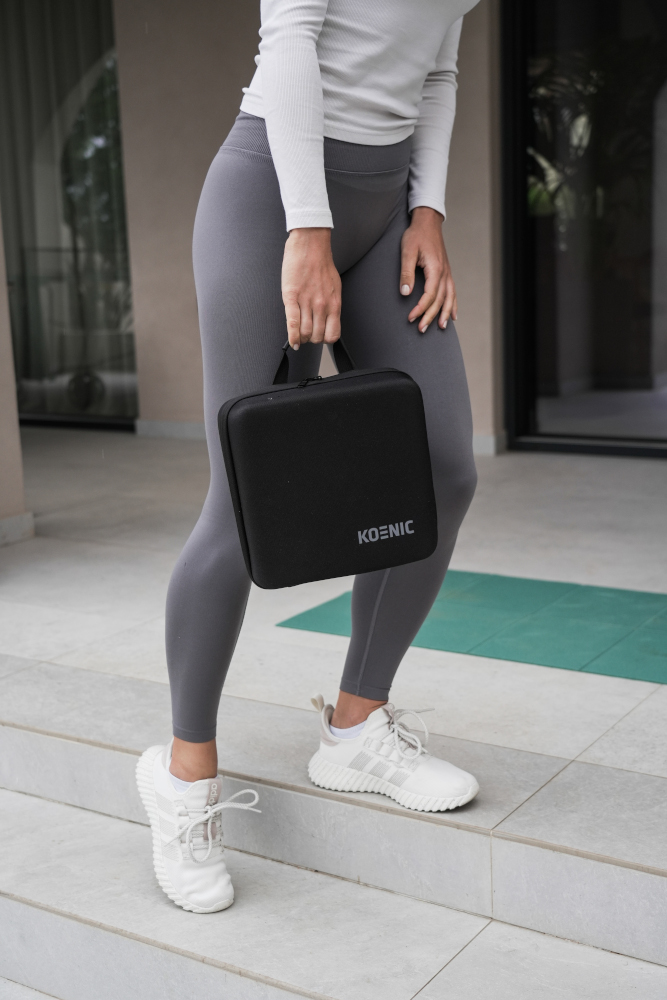 Woman in gray leggings and white shoes holding a black bag with the word 'KOENIC'.