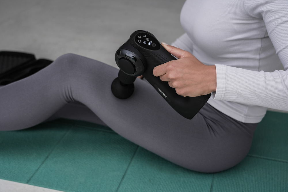 Woman using a Koznic massage gun on her leg, on a green yoga mat.