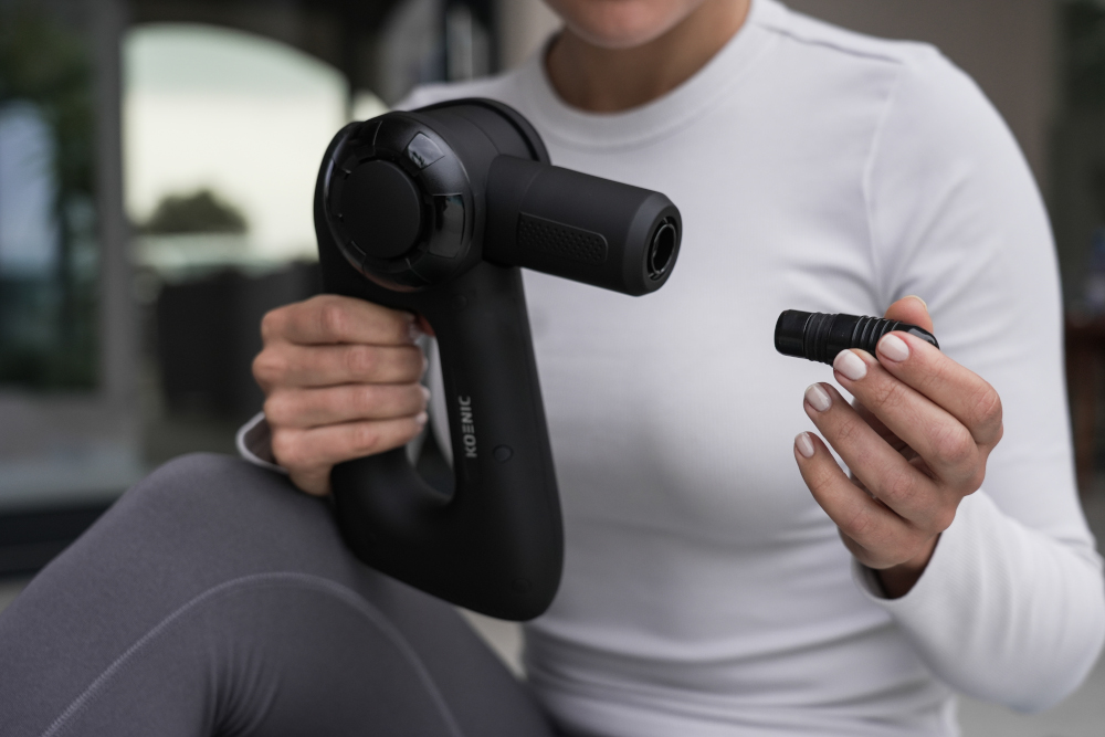 A person holding a black massage gun, attaching a head. White background.