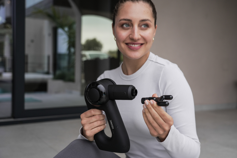 Woman holding a black massage gun and an attachment, smiling at the camera.