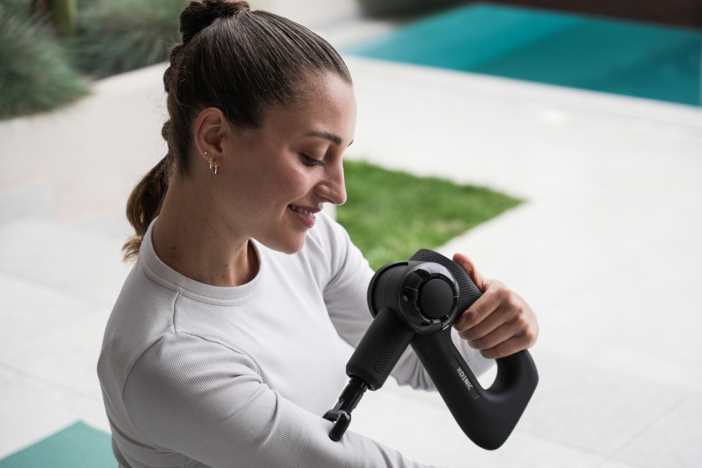 Woman using a black massage gun on her arm outdoors near a pool.