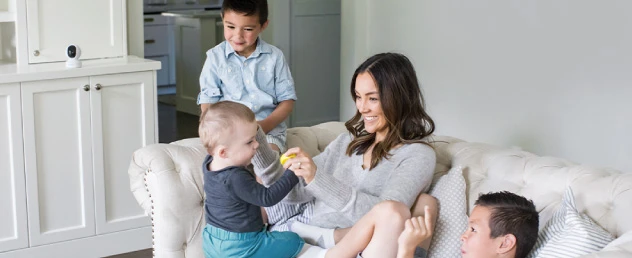 Una madre e tre bambini su un divano. Un muro bianco e un armadio. Una telecamera sull'armadio.