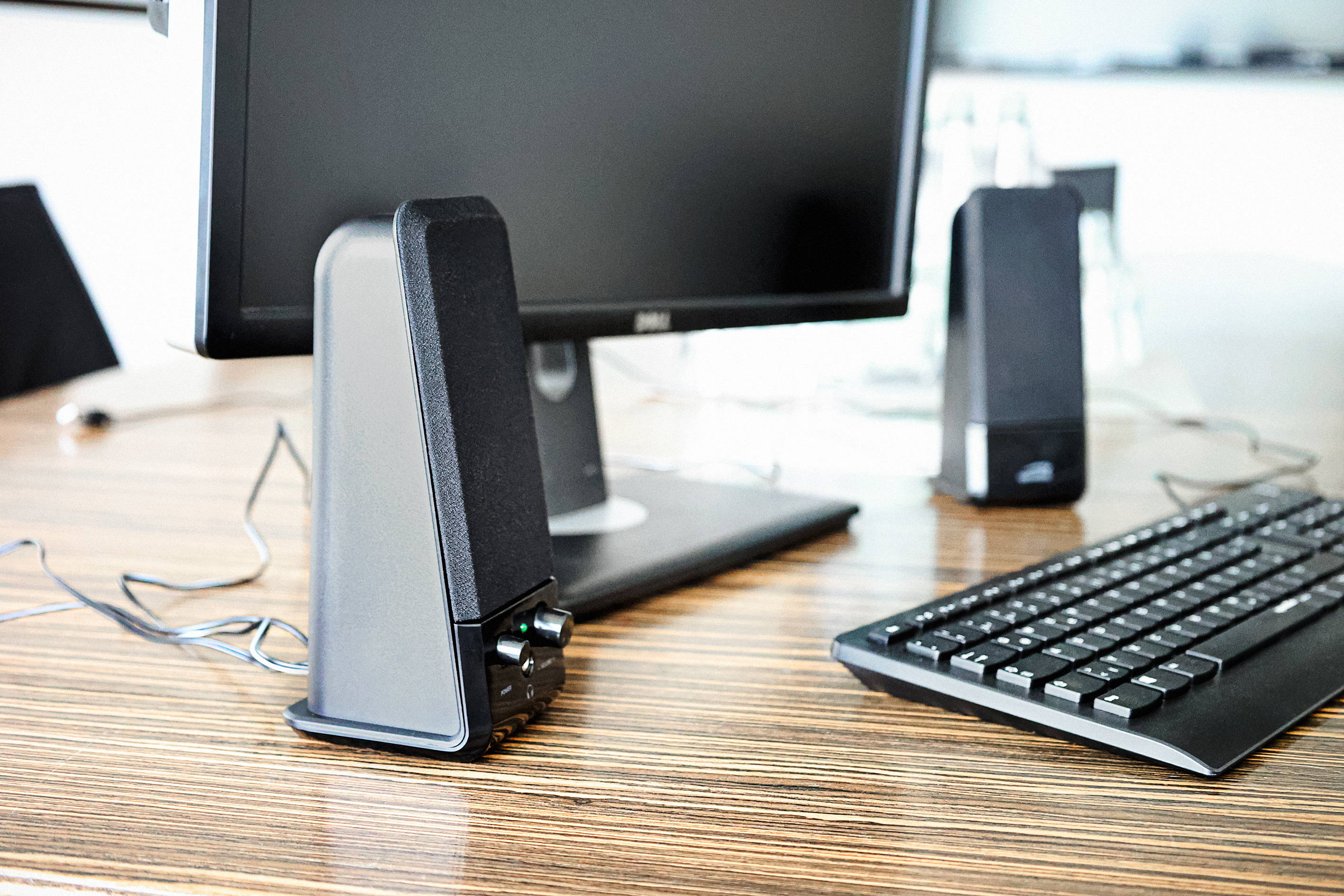 Ein Schreibtisch mit Monitor, Tastatur und zwei schwarzen Lautsprechern. Lautsprecher haben Lautstärkeregler.
