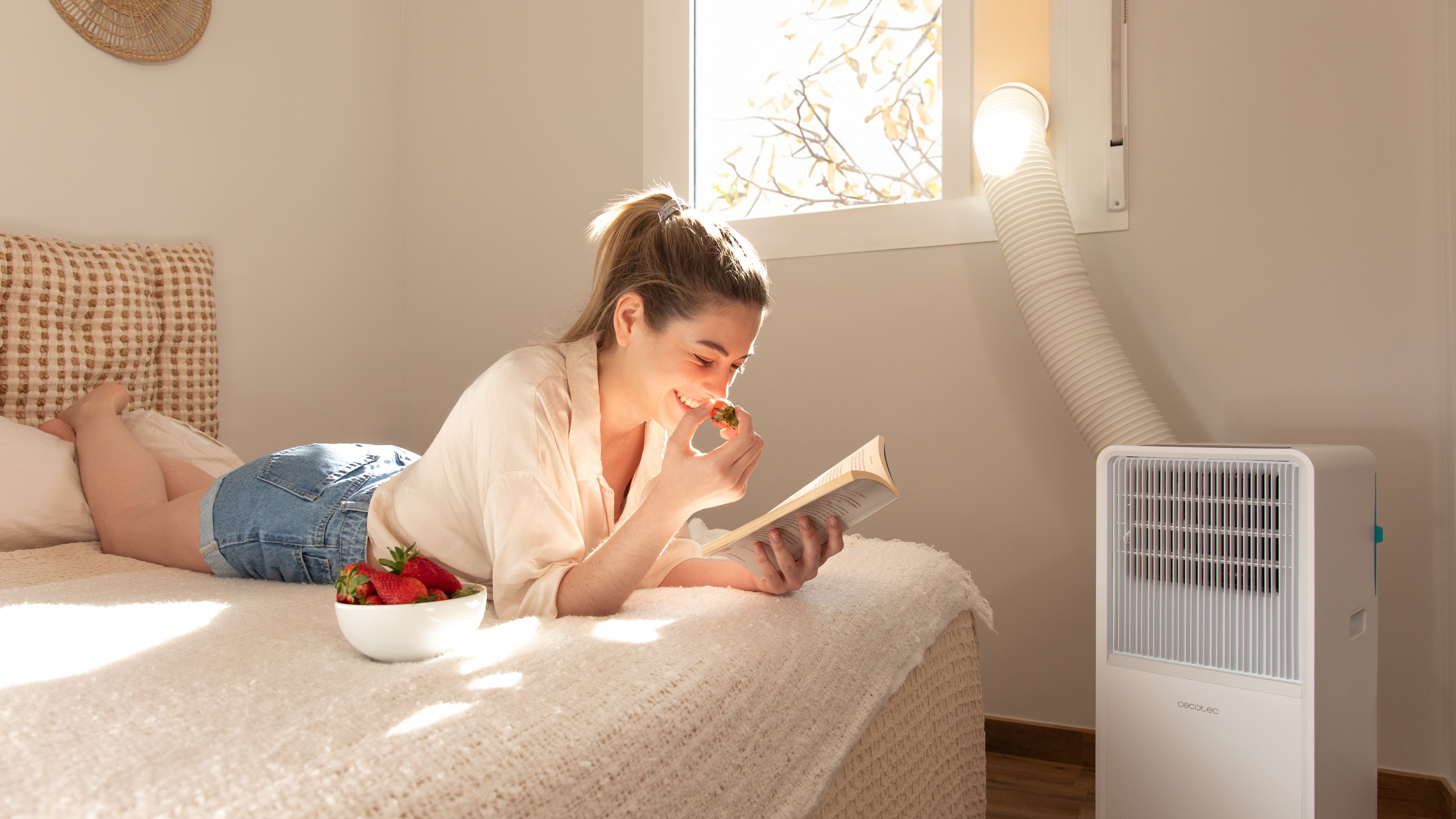 Mujer en cama leyendo libro, comiendo fresas. Aire acondicionado junto a ventana.