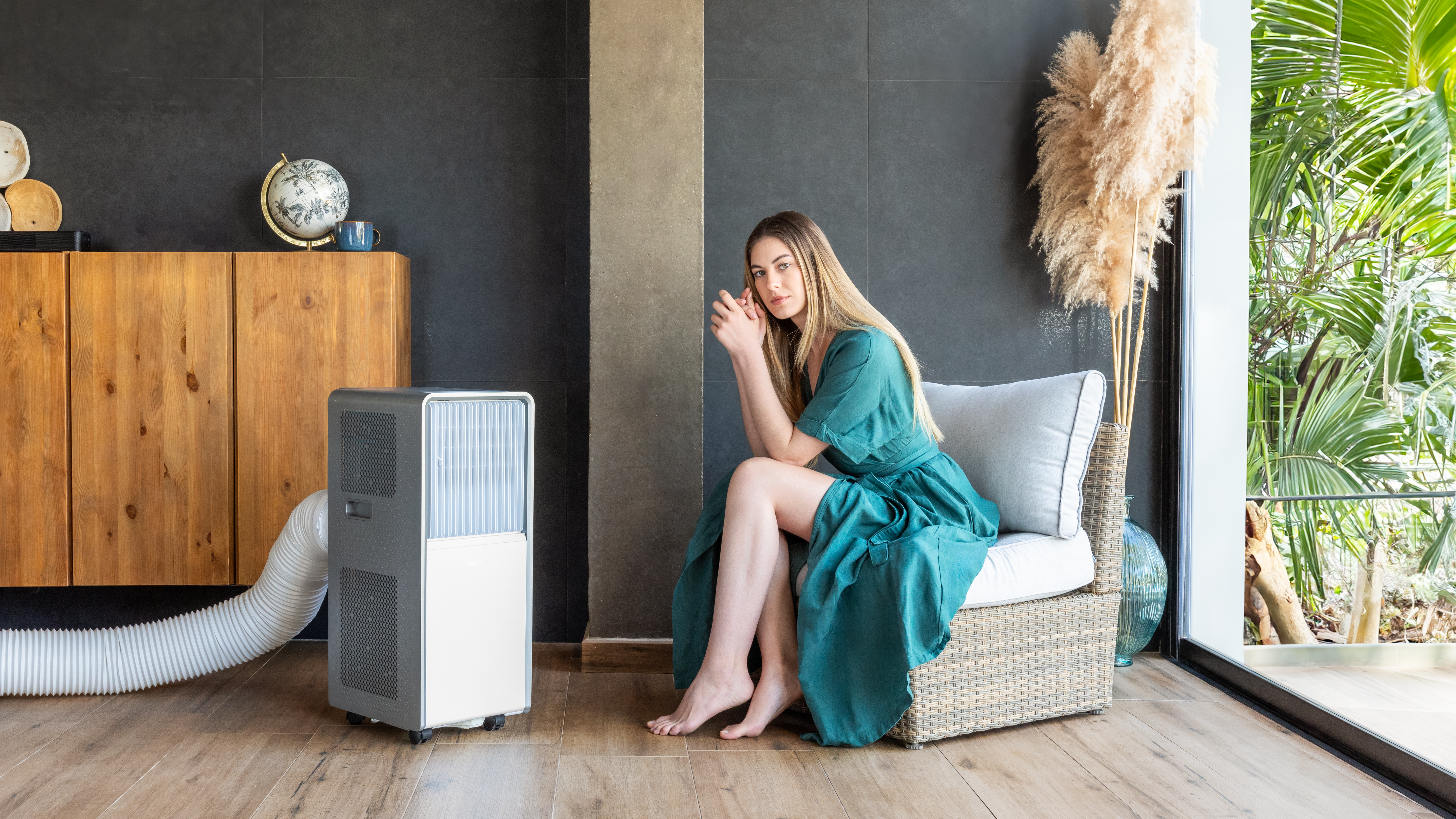 Mujer con vestido verde sentada cerca de un acondicionador de aire. Habitación con muebles de madera.