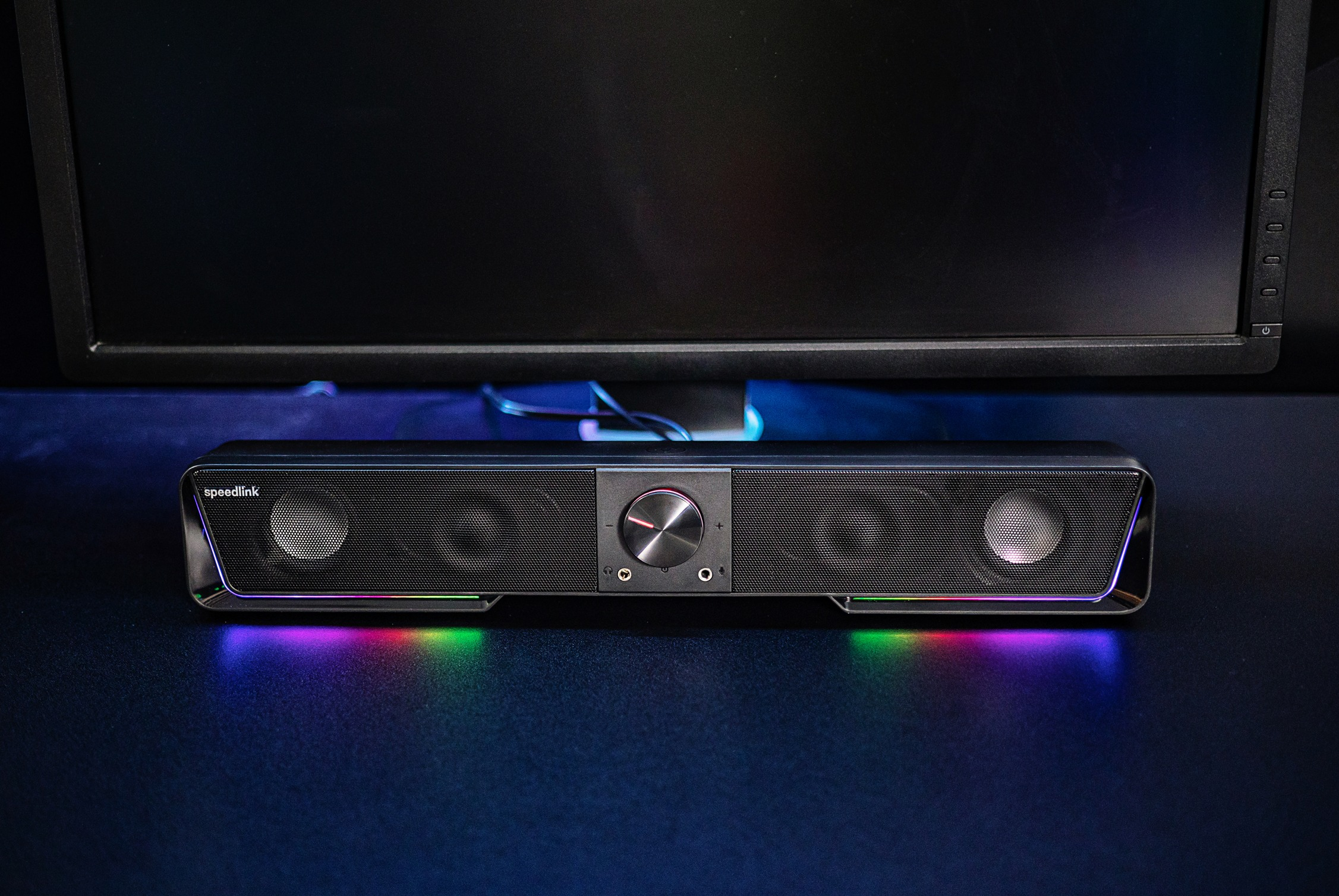 Schwarzer Soundbar-Lautsprecher mit bunten Lichtern darunter. Er steht auf dunkler Oberfläche vor Monitor.