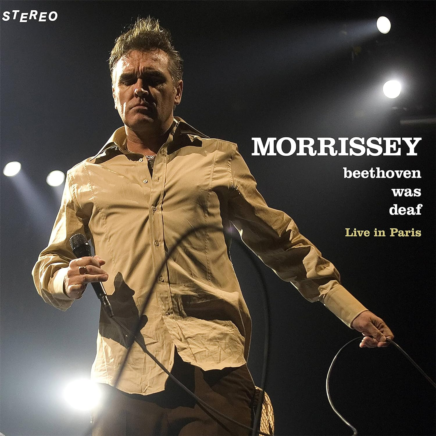 Morrissey, élő előadás. Férfi mikrofonnal, világosbarna ingben. Szövegréteg, fekete háttér.