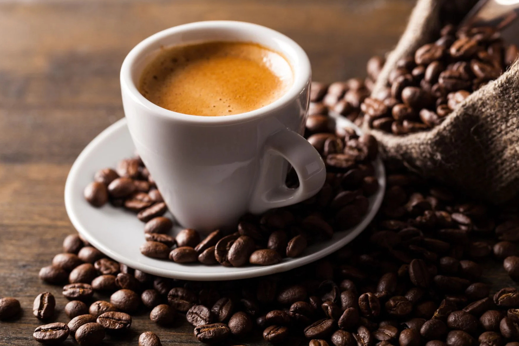 Biała filiżanka kawy na spodku otoczona ziarnami kawy, z workiem jutowym w tle.