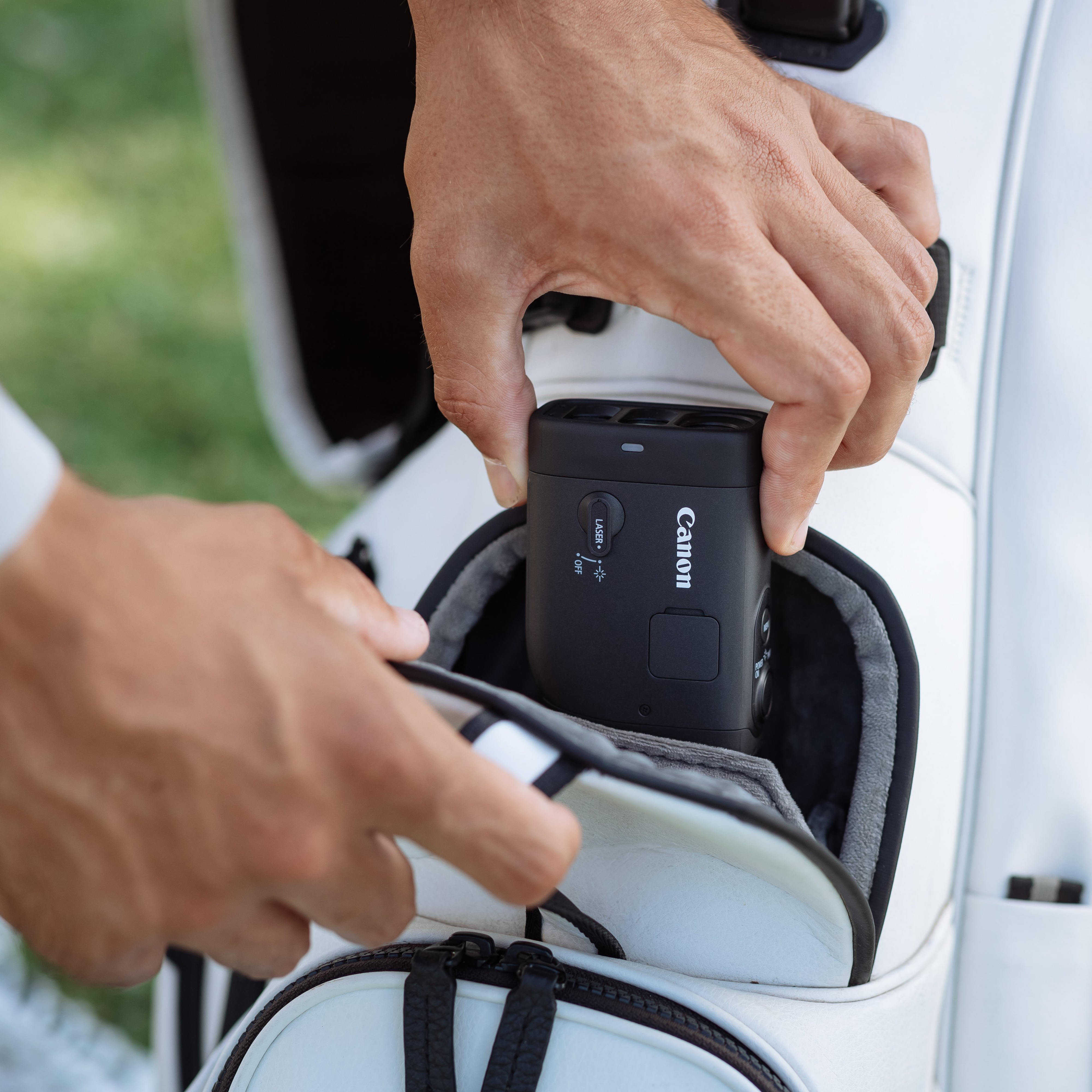 Mains plaçant un télémètre Canon noir dans un sac de golf blanc. Herbe verte en arrière-plan.