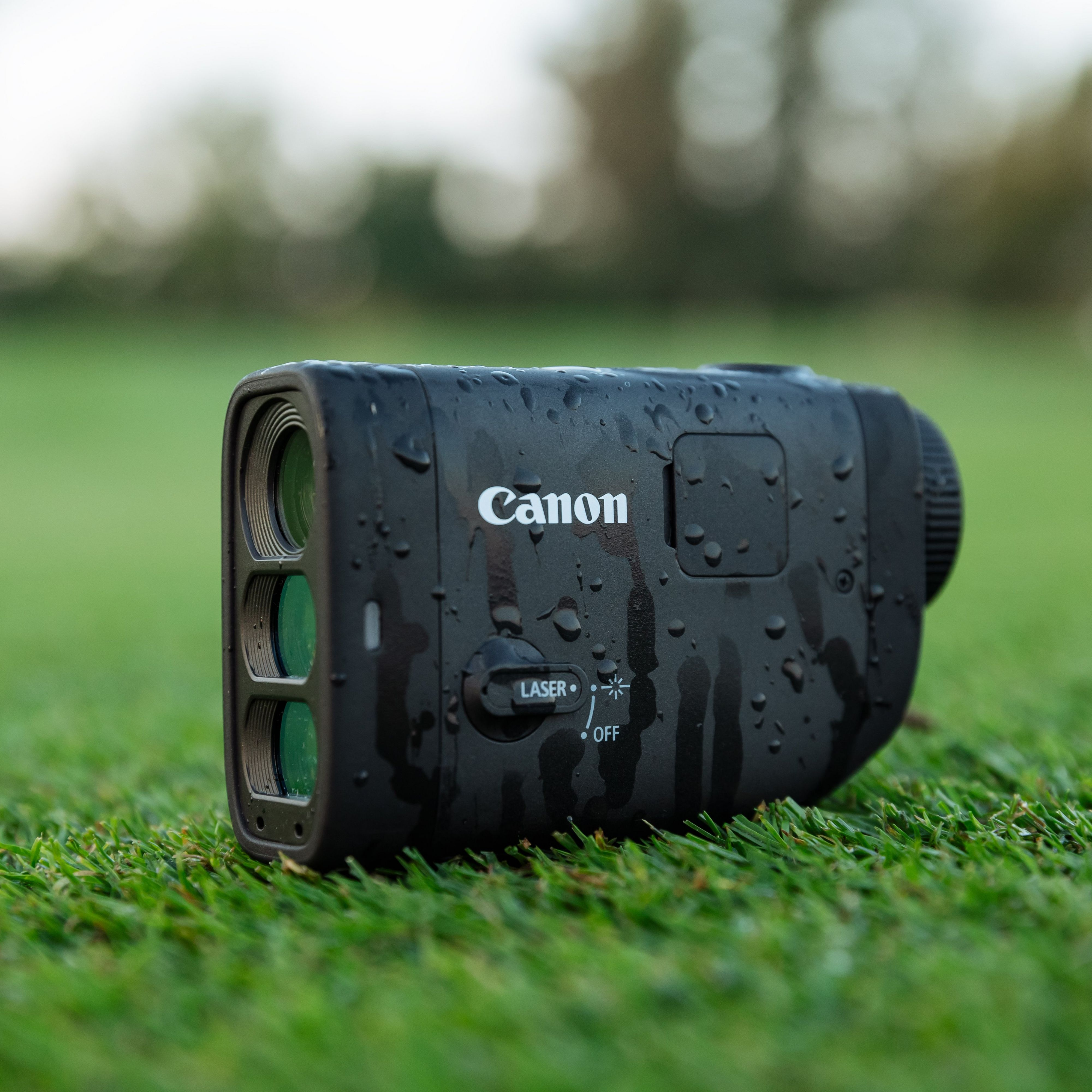 Un télémètre Canon noir avec des gouttelettes d'eau est posé sur de l'herbe verte. L'arrière-plan est vert flou et clair.