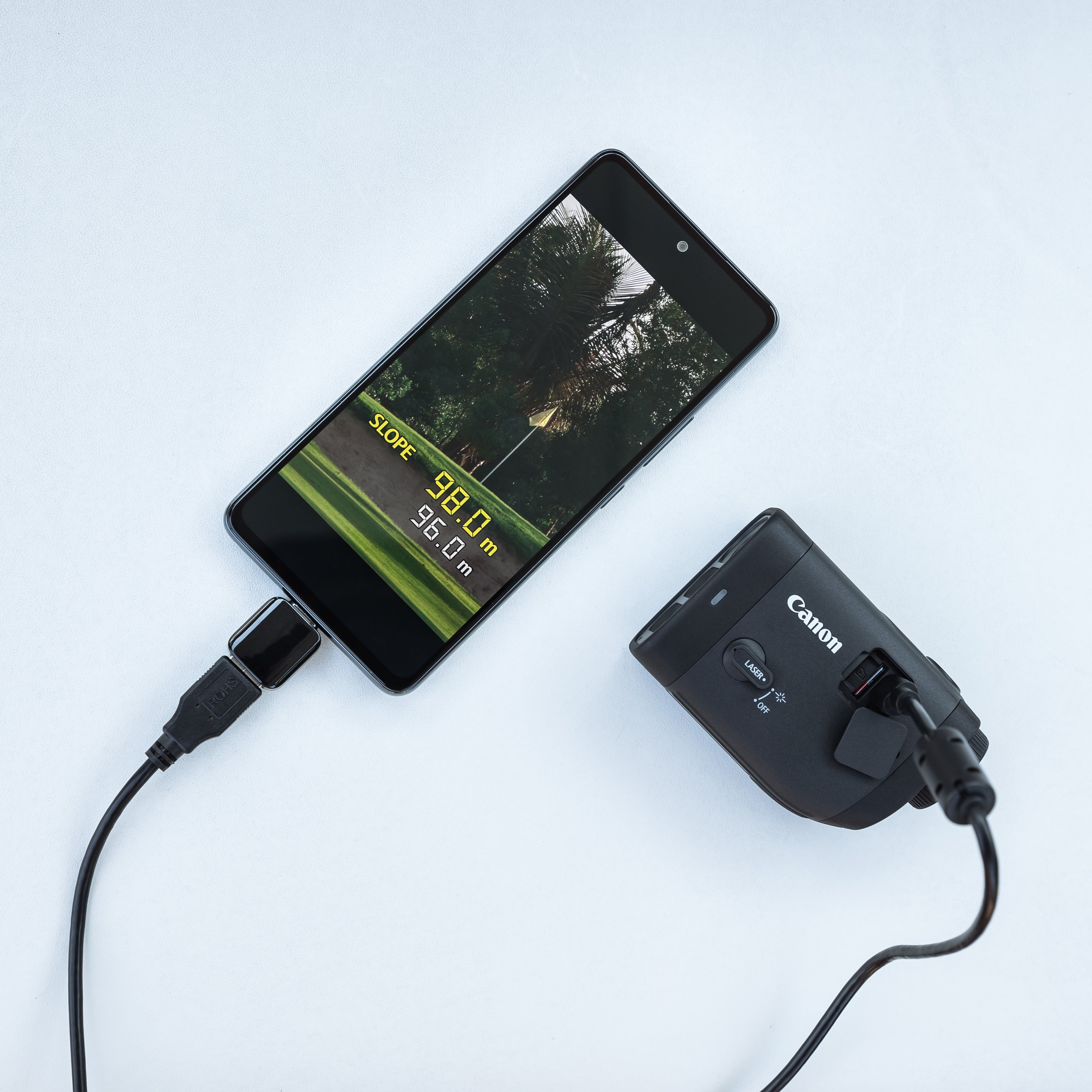 Un smartphone affiche une image de terrain de golf, connecté à un télémètre Canon. Les deux sont sur une surface blanche, reliés par un câble.