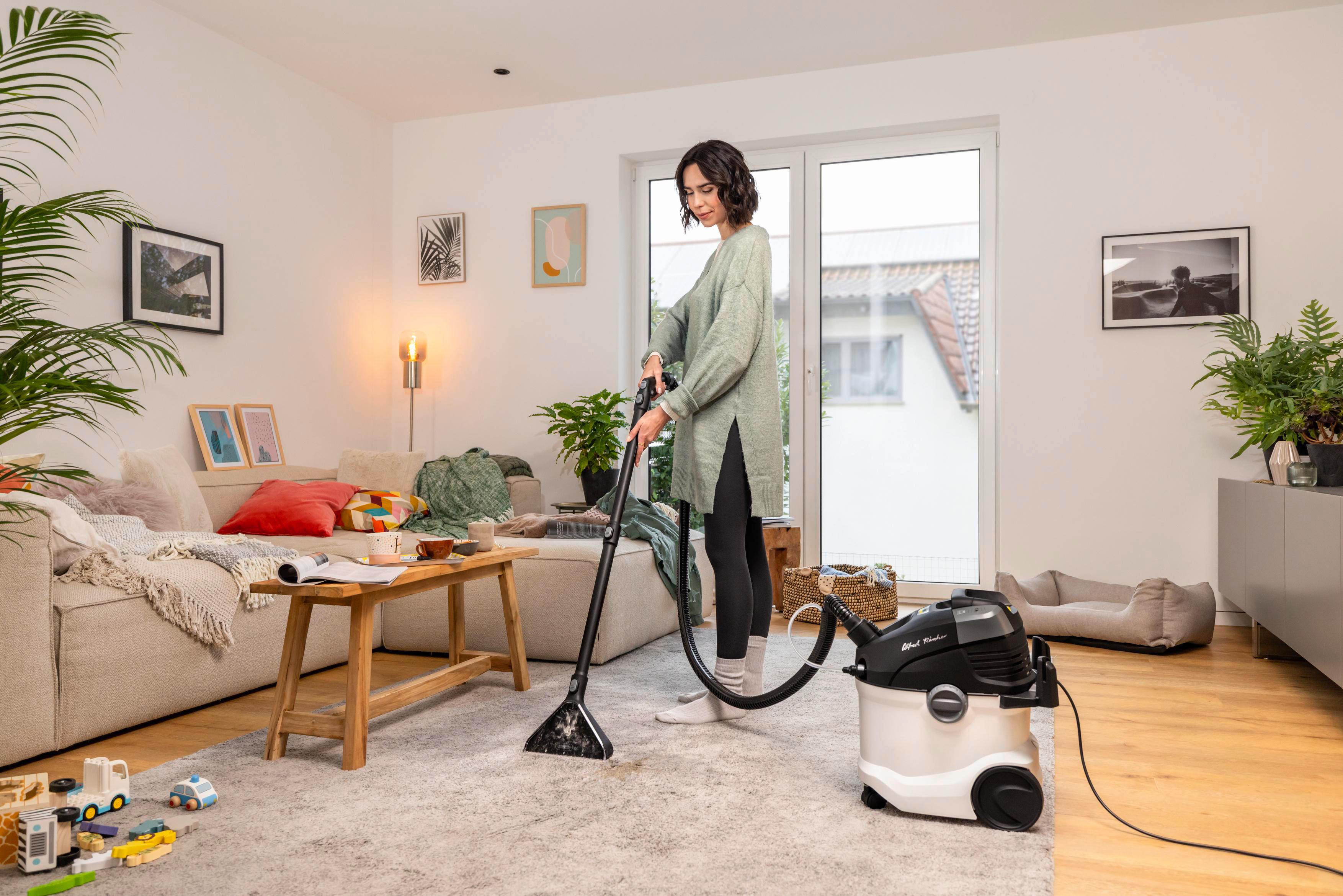 Frau, die ein Teppichwohnzimmer mit Sofa und Pflanzen staubsaugt.