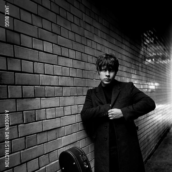 Albumhoes van Jake Bugg die tegen een bakstenen muur leunt. Hij draagt een zwarte jas en houdt een koffer vast.
